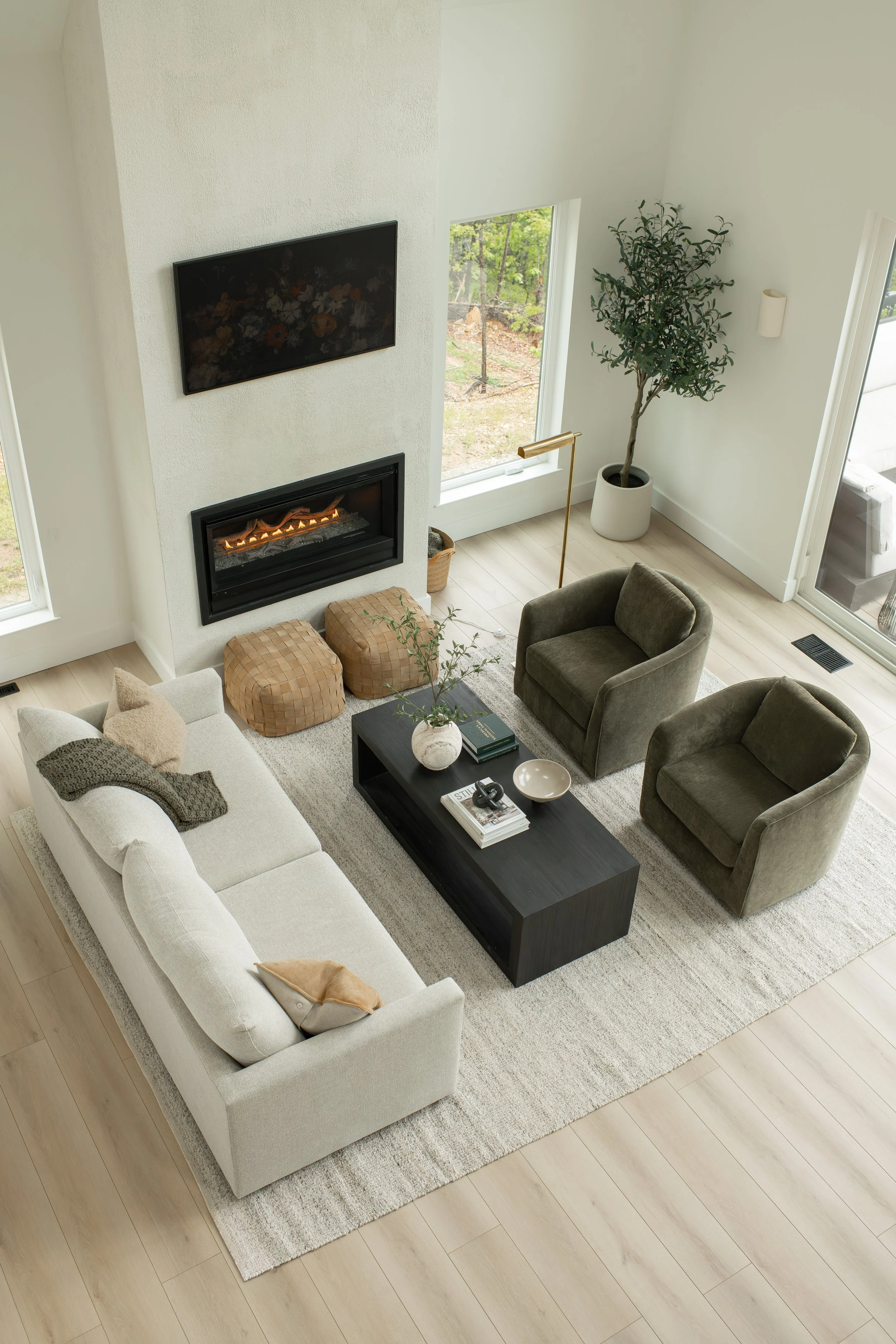 Living room with a white sofa, two brown ottomans, a black coffee table with books, a green armchair, a fireplace, a wall-mounted television, large windows with a view of outdoor greenery, a potted plant, and a tall floor lamp.