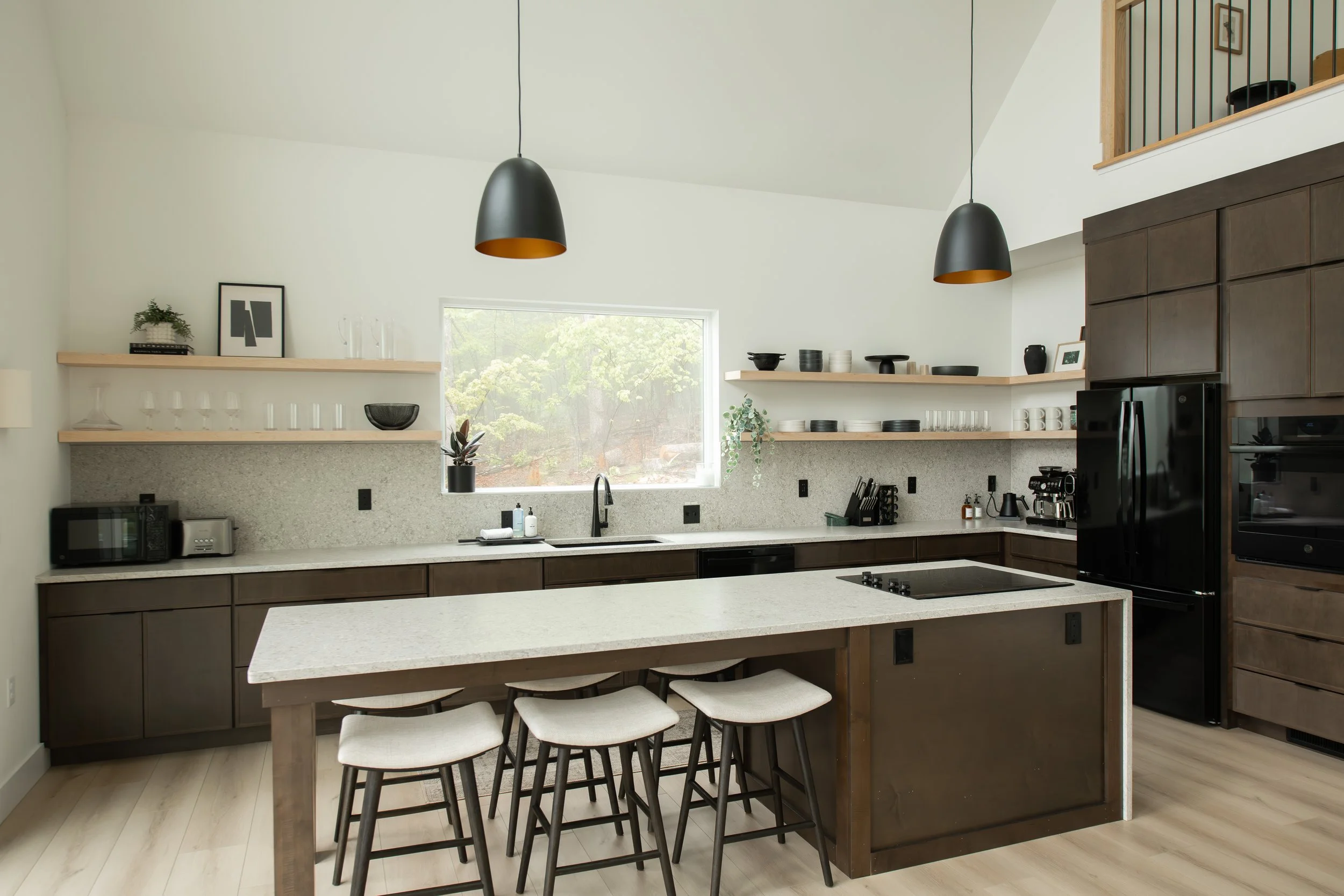 Modern kitchen with a beige island and dark brown cabinets, open shelves with dishes, black pendant lights, and a window overlooking trees.