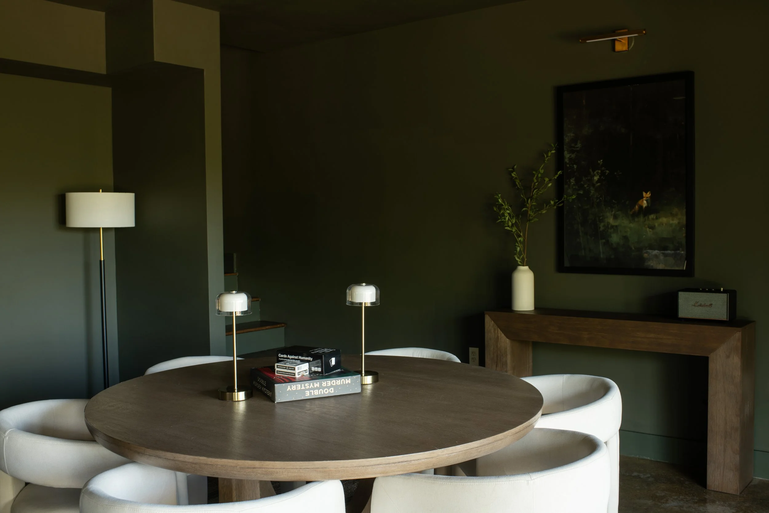 A modern dining area with a round wooden table surrounded by white chairs, a marble lamp, a white vase with green branches, a framed painting of a fox, and a bookshelf with books.