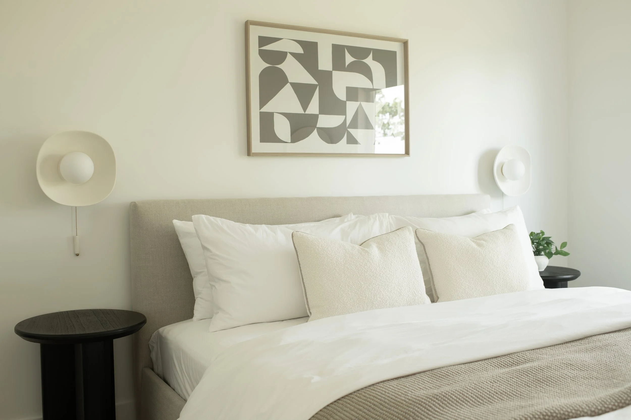 Modern bedroom with white bedding, beige headboard, abstract art painting above bed, white wall sconces, black side table, and green plant.