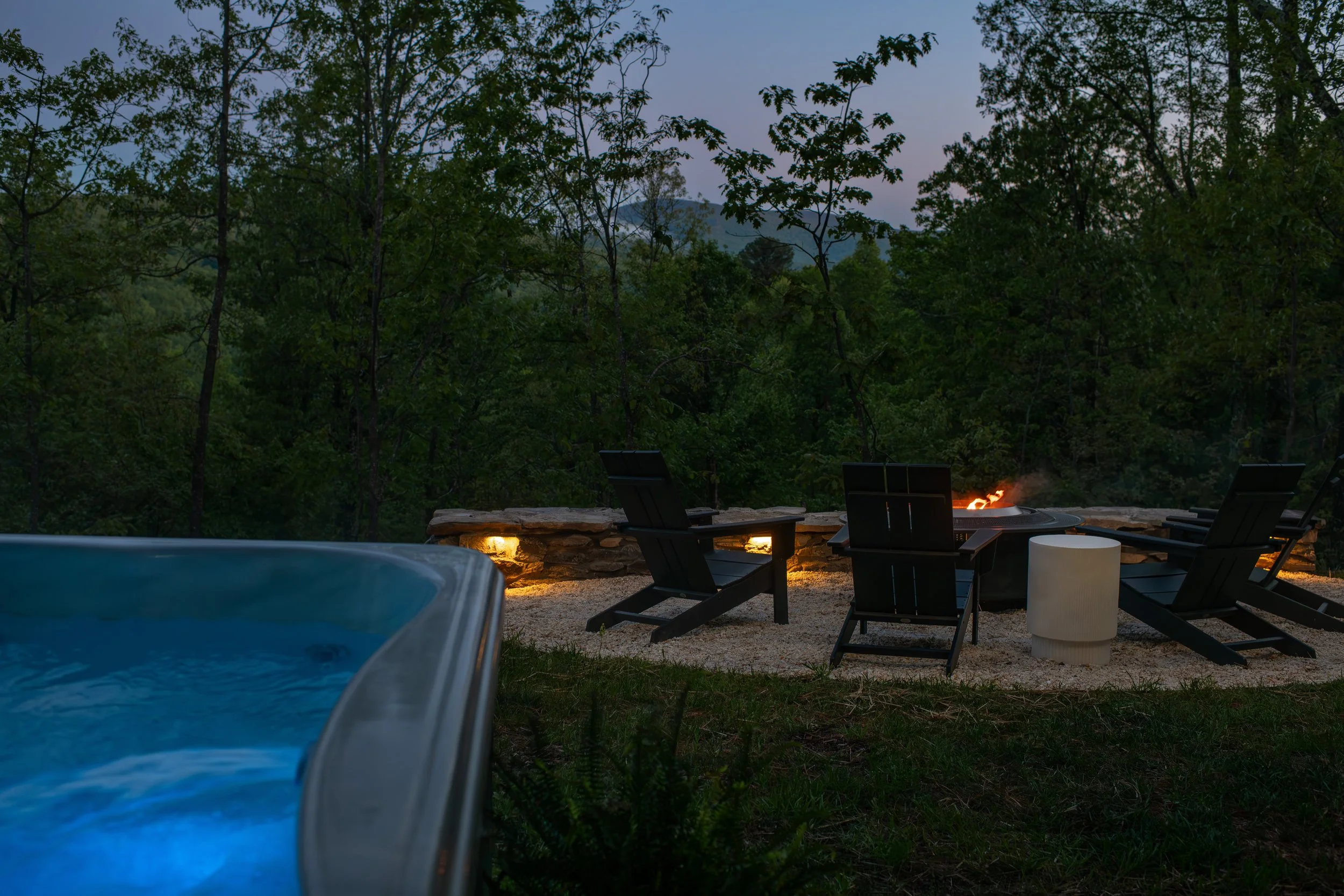 Outdoor patio with lounge chairs and fire pit, surrounded by trees, during early evening.