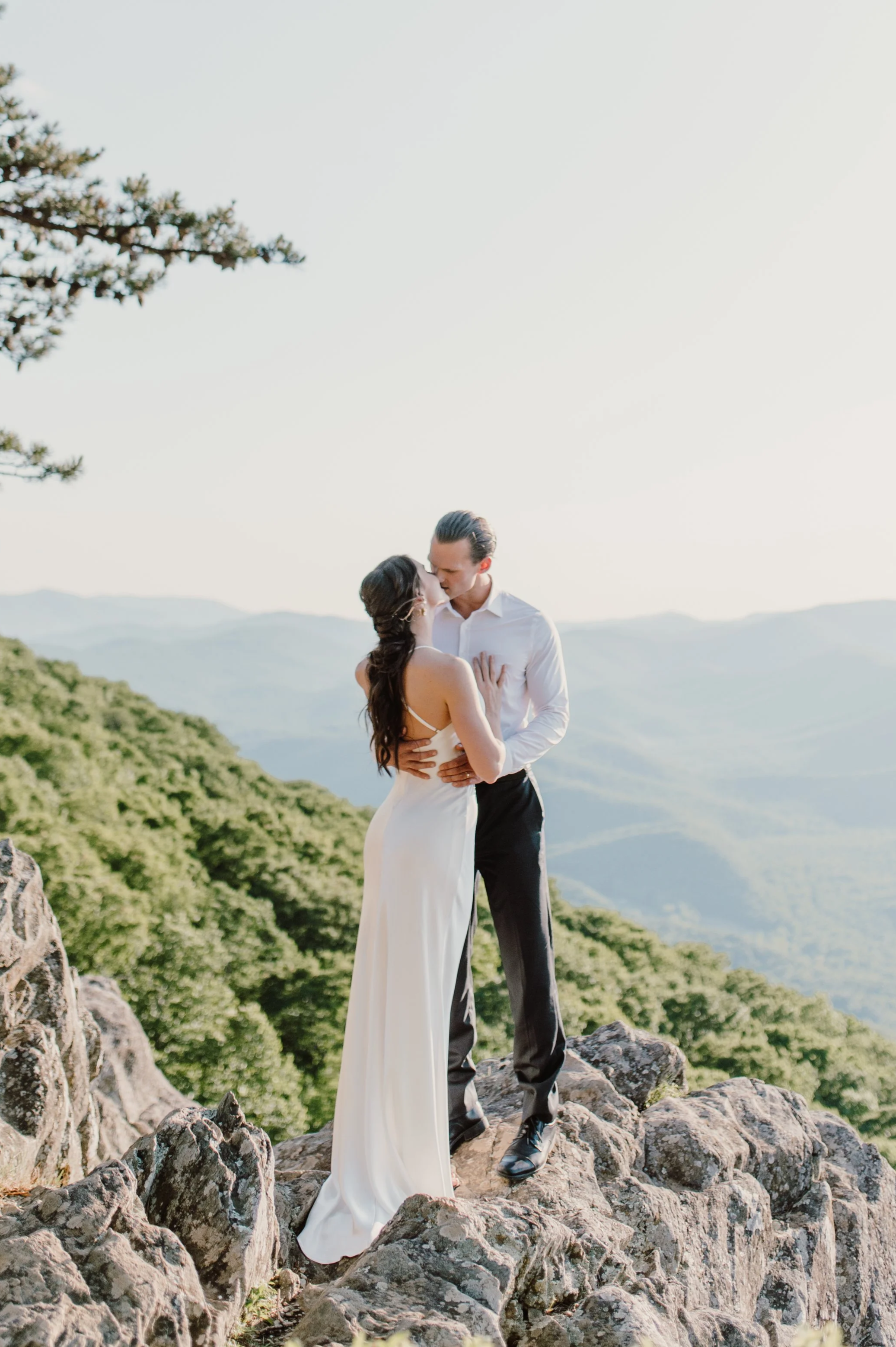 A couple in wedding attire standing on rocks outdoors with mountains and a clear sky in the background, sharing a kiss.