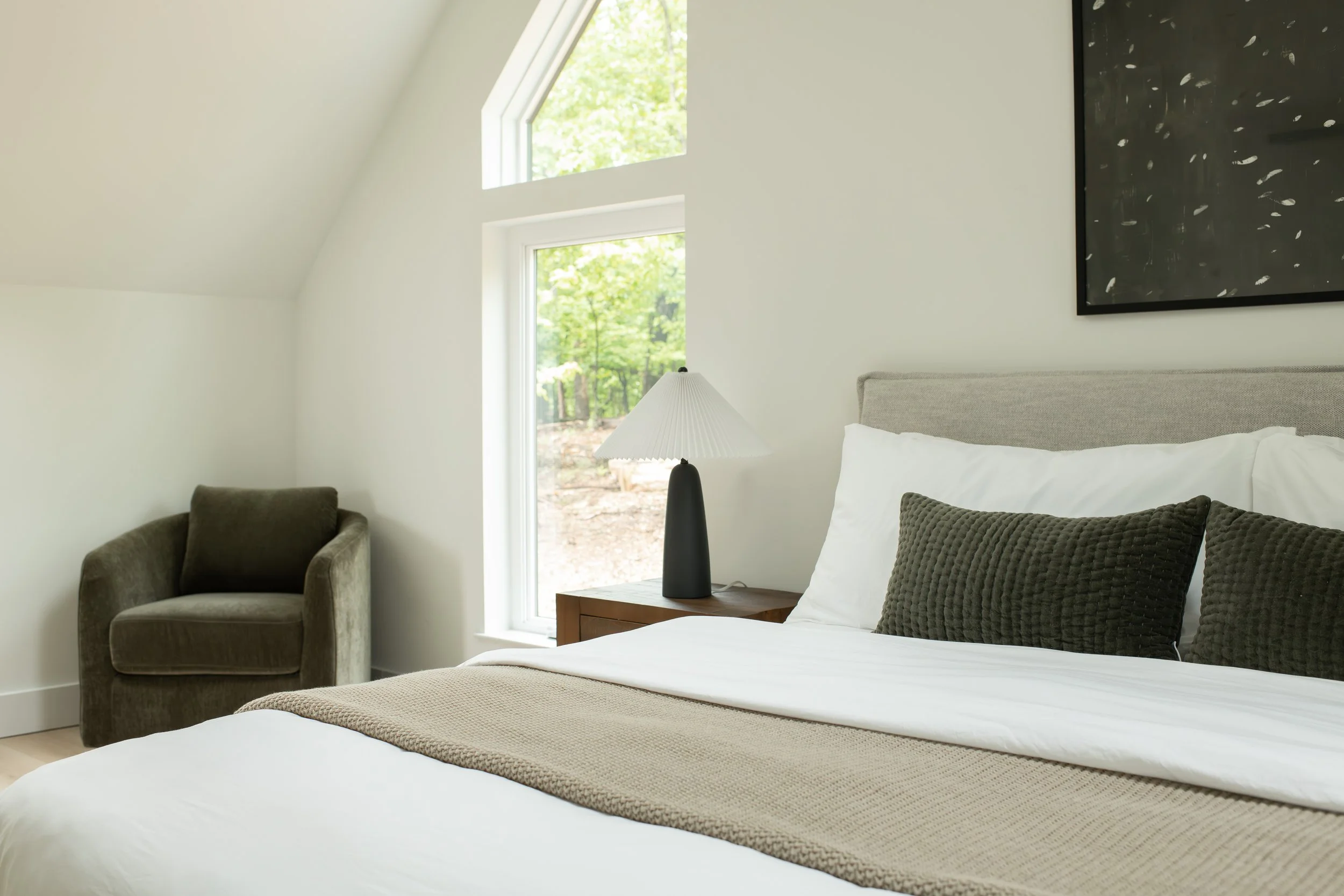 A bedroom with white walls and a sloped ceiling, featuring a large window with greenery outside. The bed has white bedding and darker accent pillows, with a black bedside lamp on a wooden nightstand. There is a dark armchair in the corner and a piece