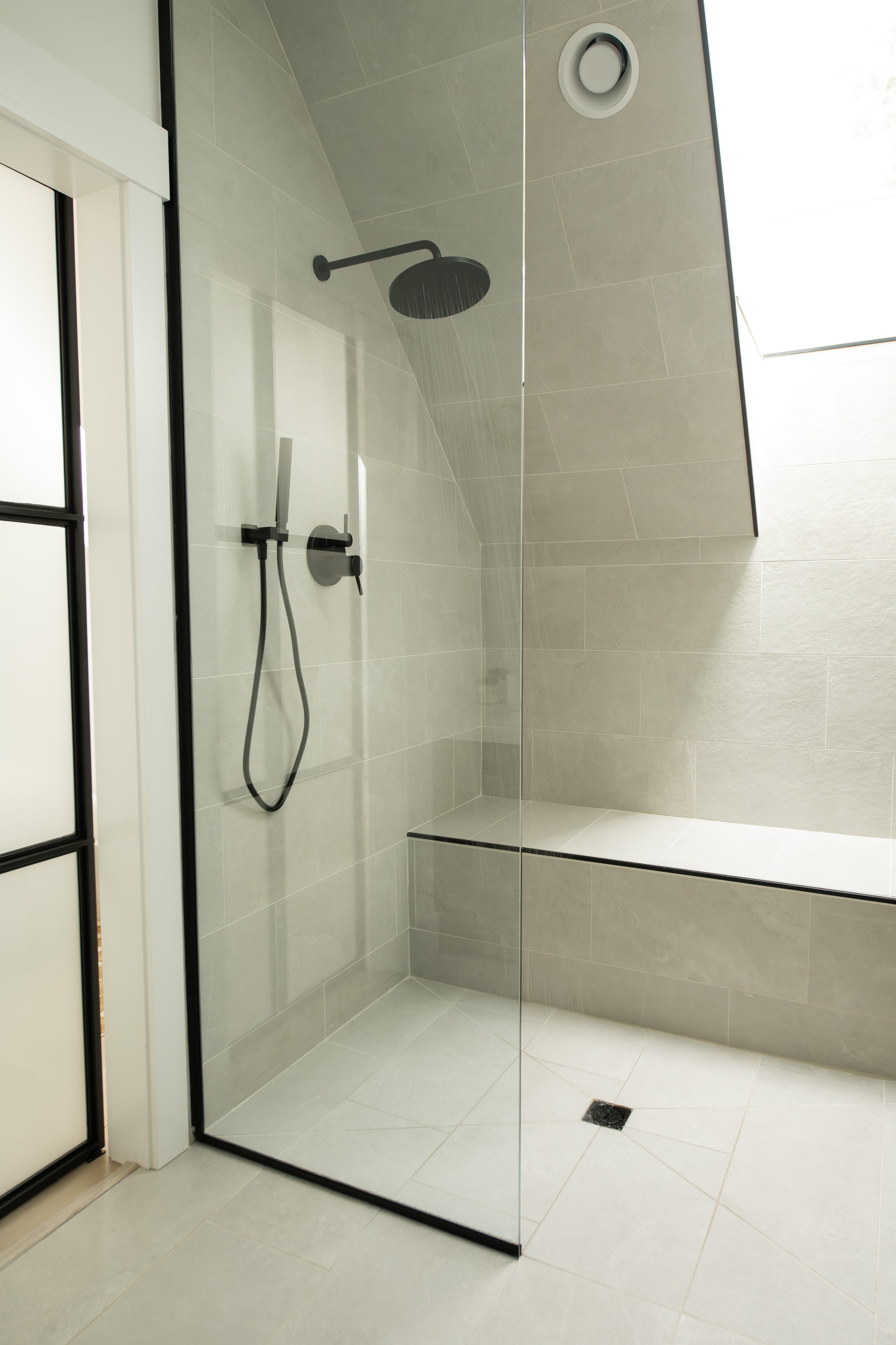 Modern walk-in shower with a black rainfall showerhead, a handheld shower, and a sleek tile wall. The shower has a glass enclosure and a built-in bench.