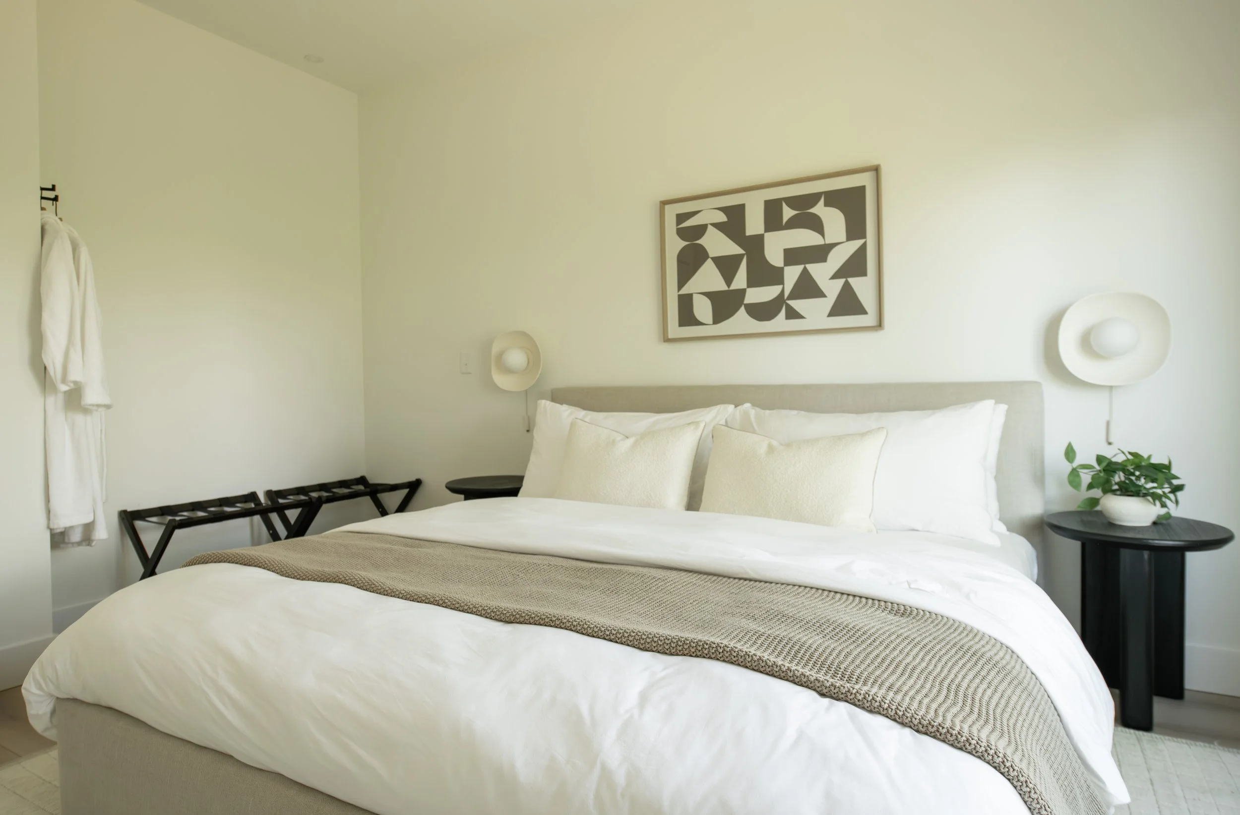 A neatly made bed with white pillows and a beige blanket in a minimalist bedroom. On the wall above the bed, there's a framed black and white abstract art piece. To the right, there's a black bedside table with a potted green plant. On either side of