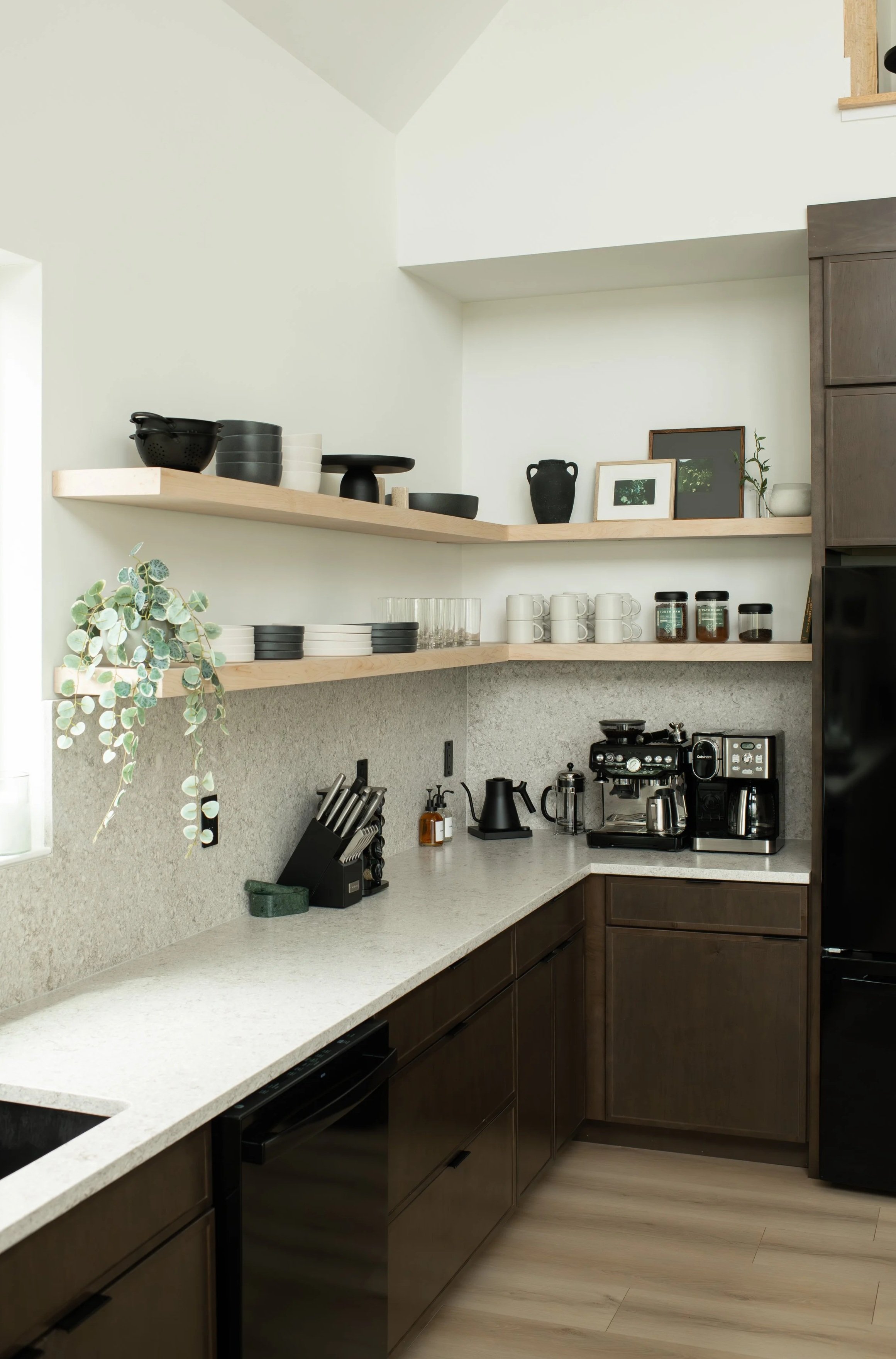 Modern kitchen with beige countertops, dark brown cabinets, open wooden shelves holding dishes and decorative items, and coffee appliances on the counter.