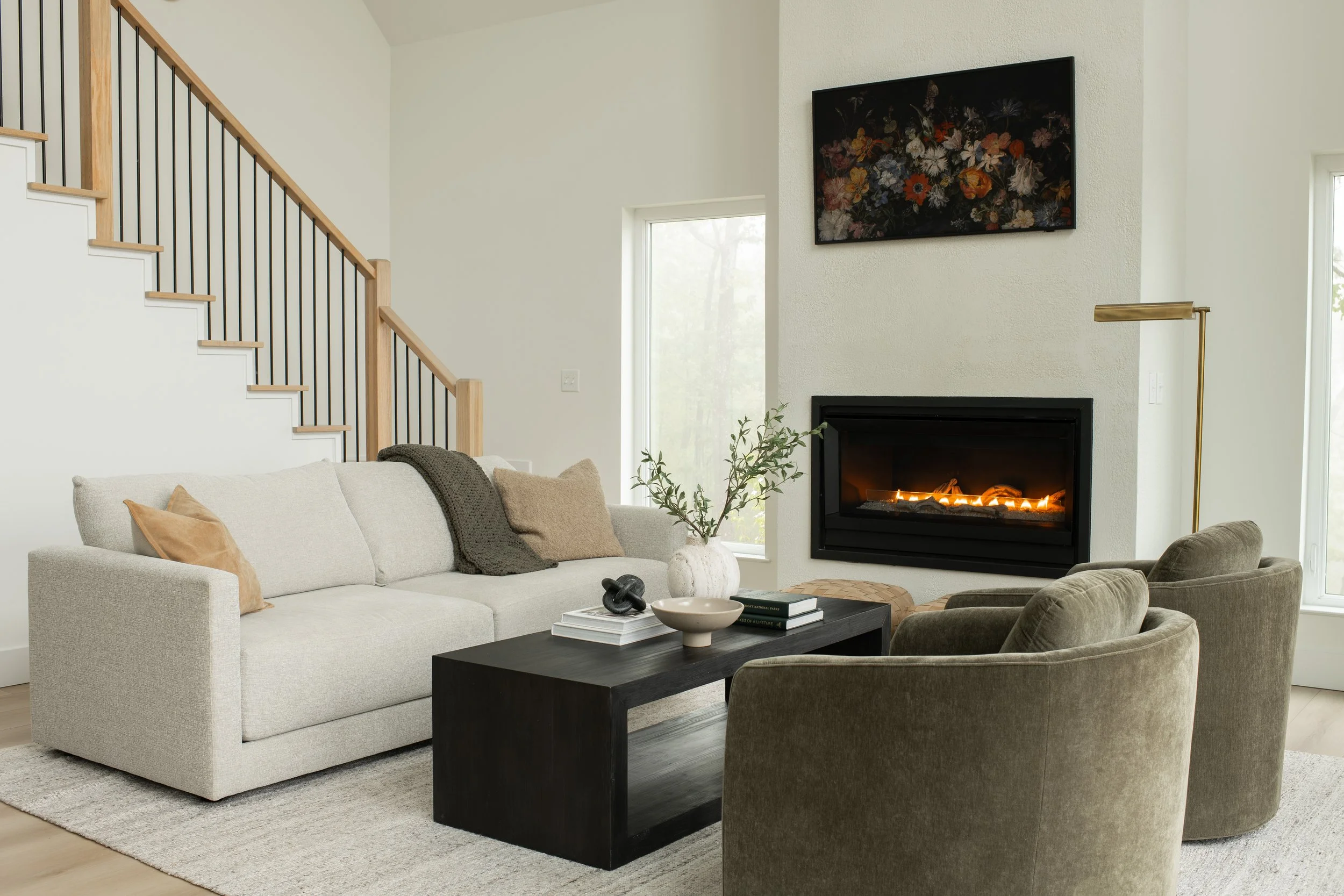 A living room with a beige sofa and dark green armchair, a black coffee table with books and decorative objects, a white textured wall with a rectangular floral painting, a fireplace with a warm fire, and large windows providing natural light.