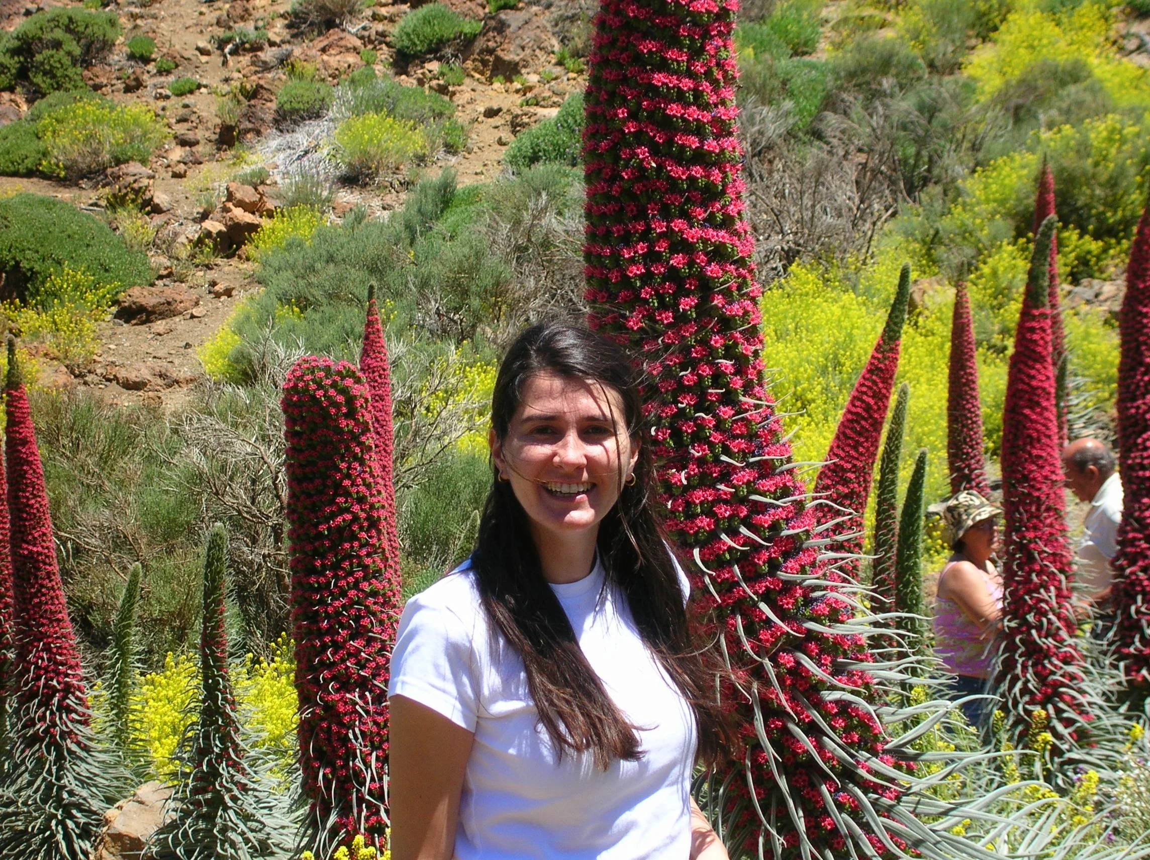A woman with long dark hair smiling in front of the spectacular red Tajinaste flowers in Tenerife, with rocky volcanic terrain in the background.