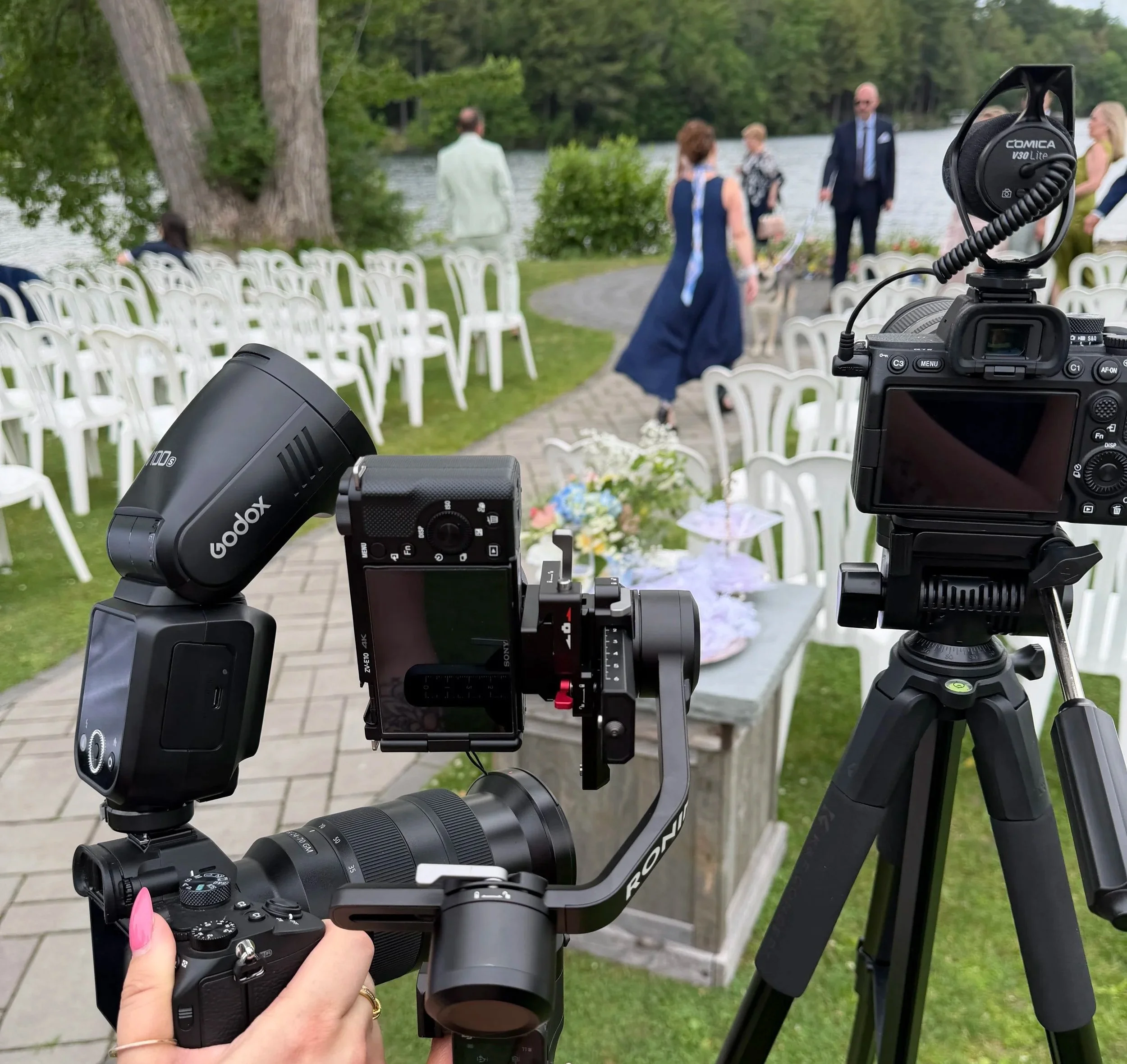 A professional camera used for filming or photography set up outdoors near a river, with a scenic background of trees and chairs arranged for an event, possibly a wedding or outdoor gathering, during daytime.
