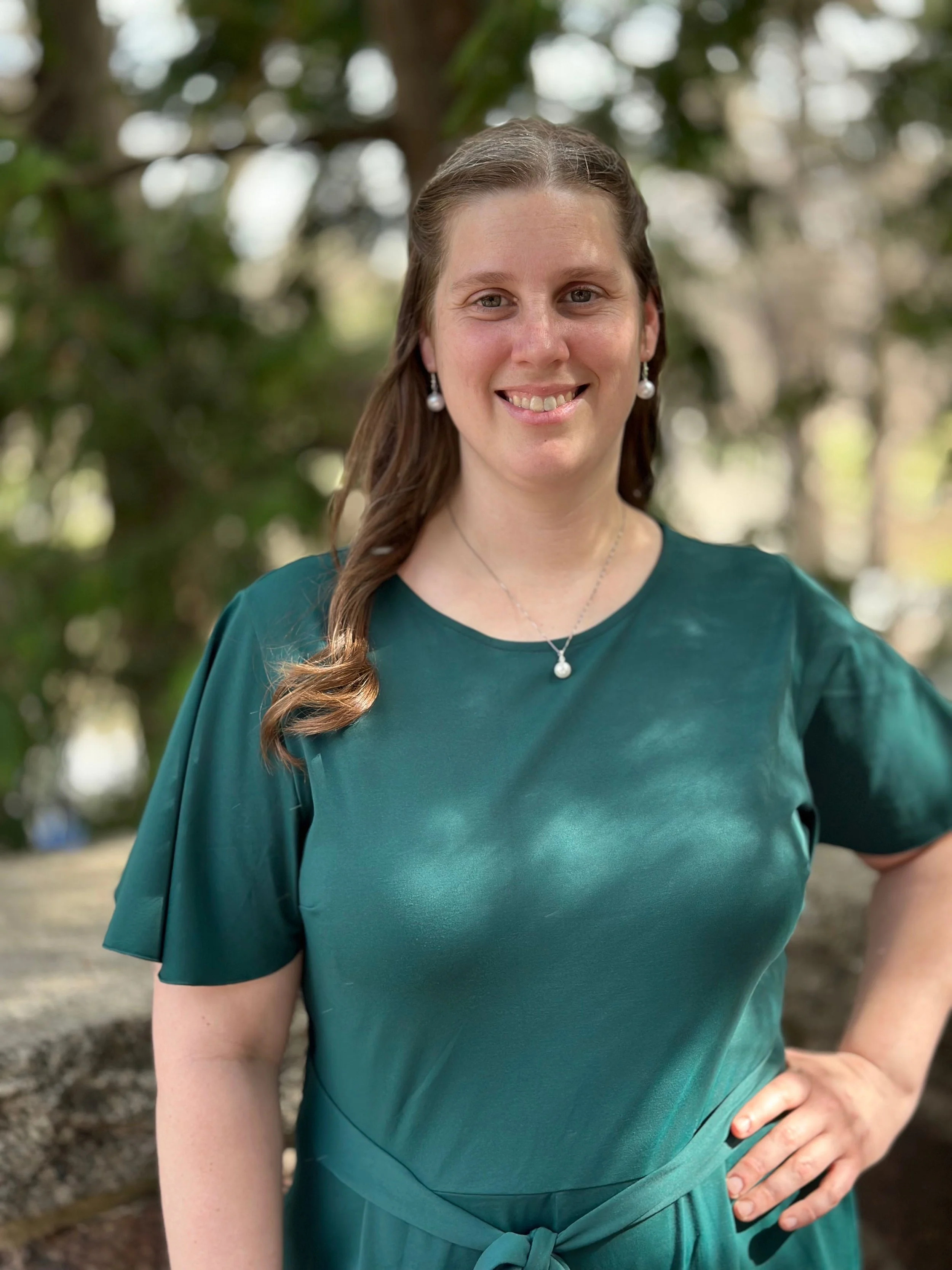 A woman wearing a teal dress with puffed sleeves, pearl earrings, and a pearl necklace, standing outdoors in front of blurred trees, smiling with her right hand on her hip.