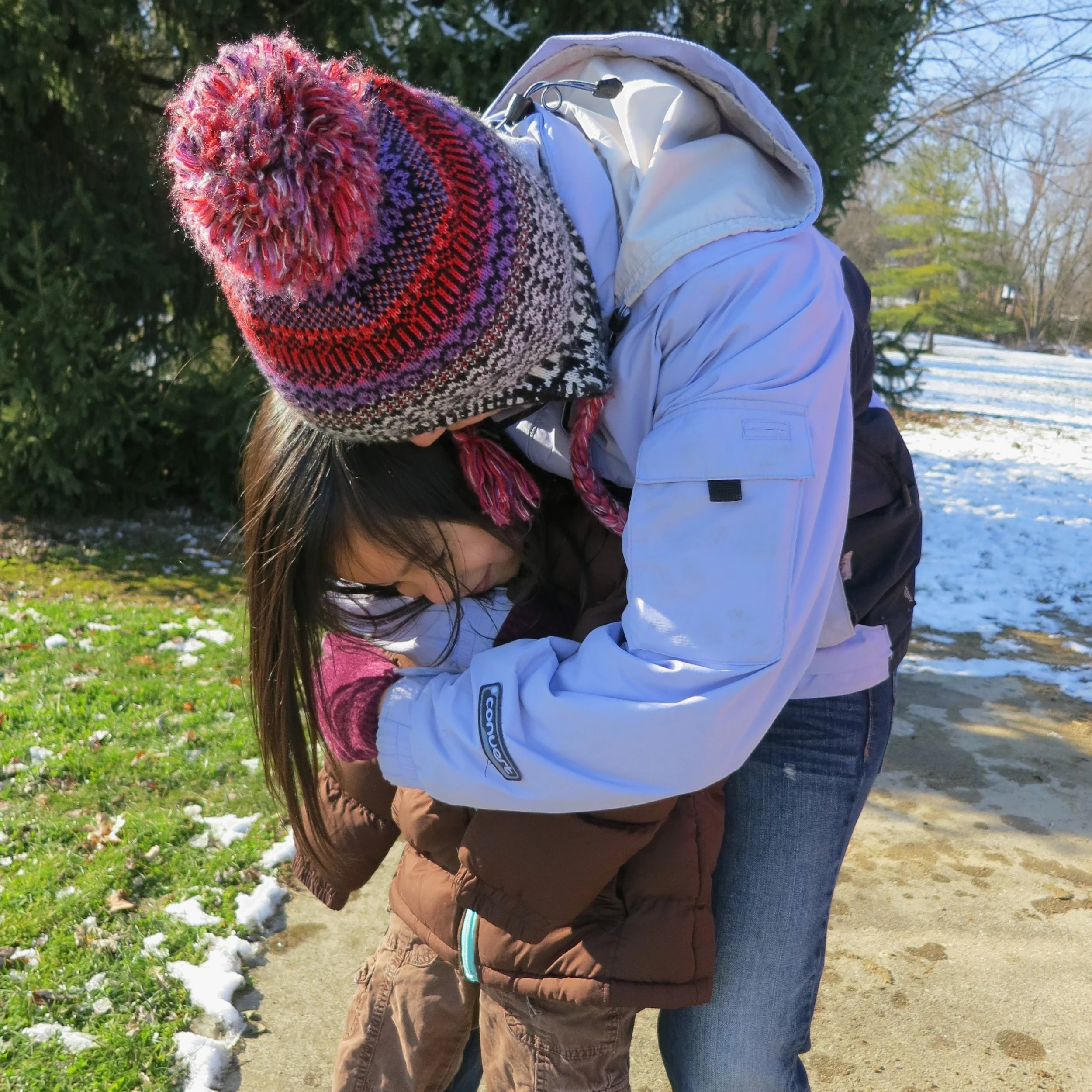A young woman wearing a light blue jacket and a colorful knitted hat with a pom-pom, leans over from behind to hug her niece, a young girl with long dark hair . The girl is receiving the hug but not embracing back. They are outdoors in the snow.