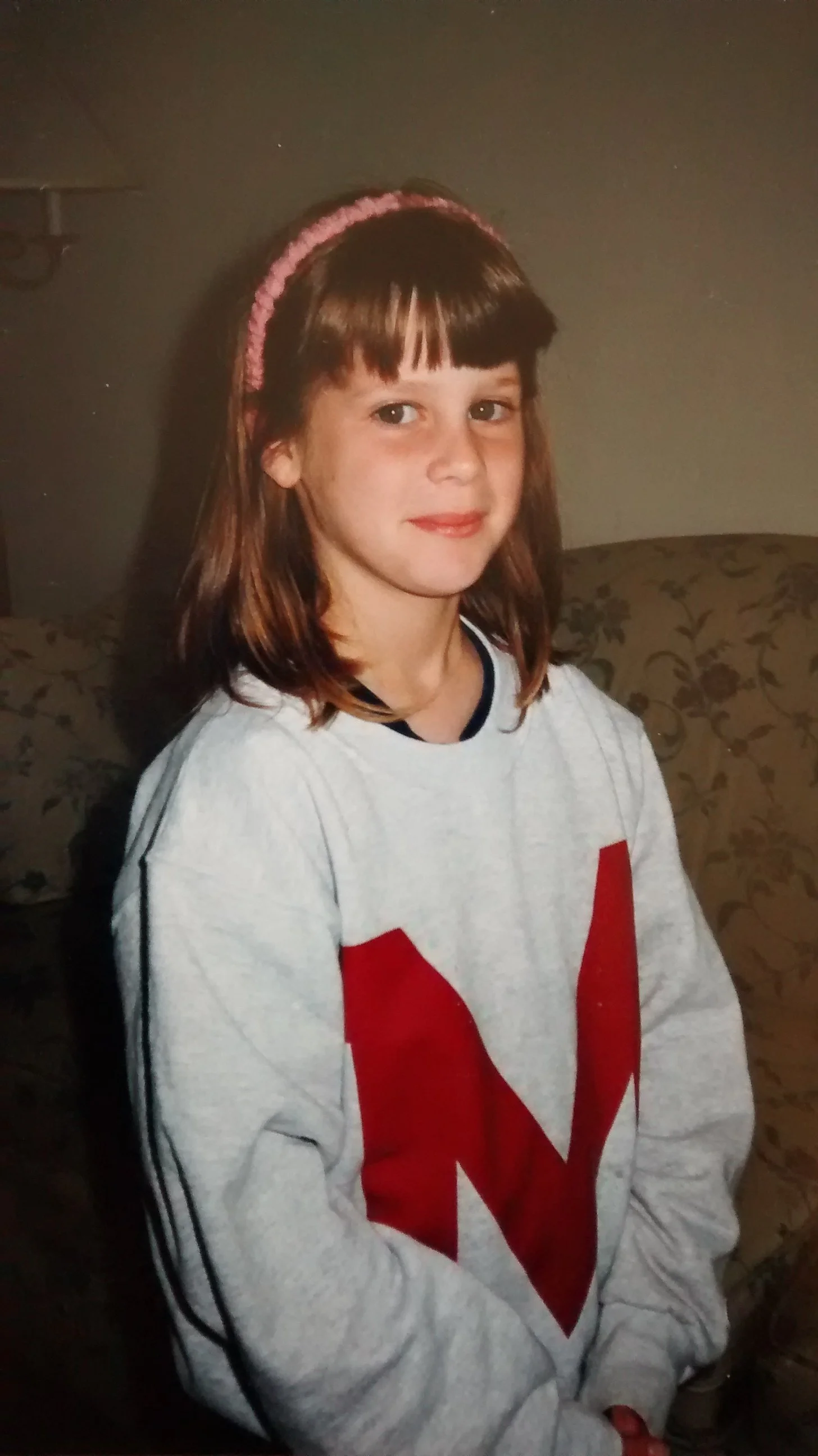 A young Annie Chase with brown hair and bangs, wearing a pink headband and a white sweatshirt with a large red letter 'M' on the front, sitting on a patterned sofa in a dimly lit room.