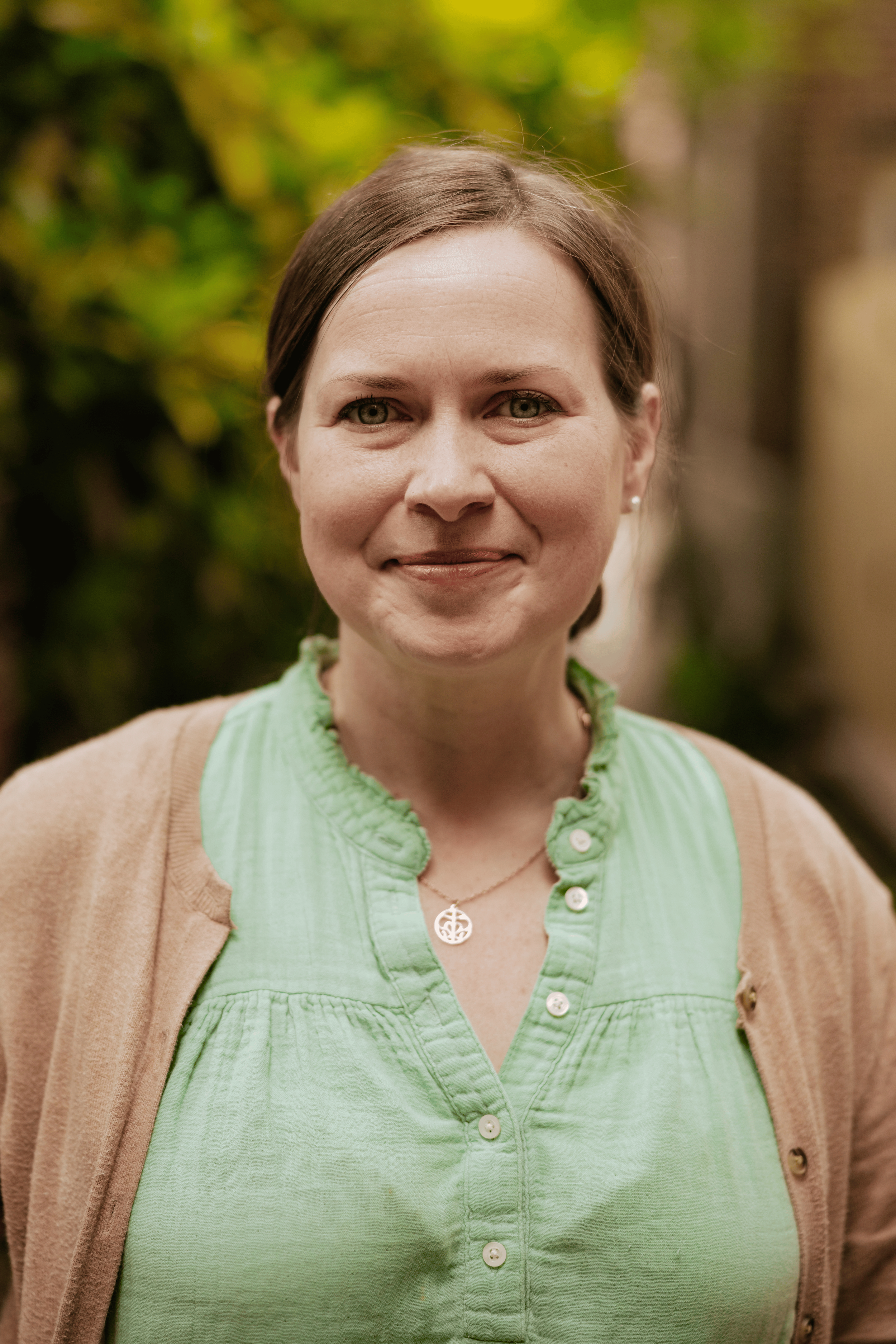 A woman with shoulder-length brown hair smiling, wearing a light green button-up shirt and a beige cardigan, standing outdoors with green foliage in the background.
