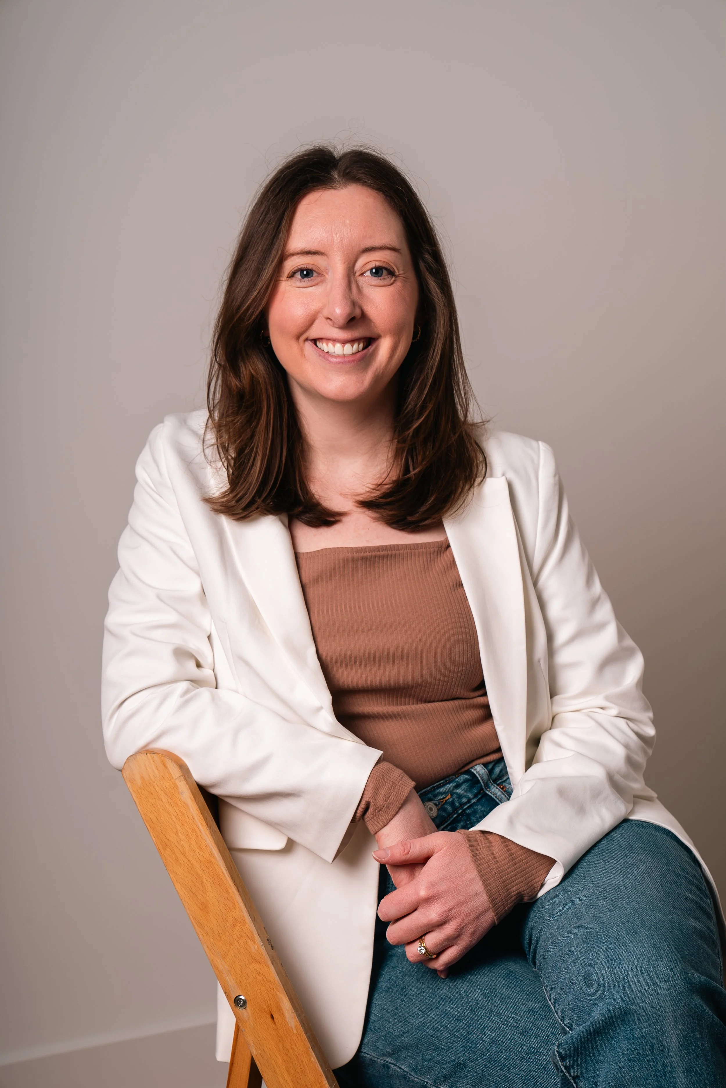 A woman with shoulder-length brown hair, wearing a white blazer over a brown top, sitting on a wooden chair against a plain gray background, smiling at the camera.