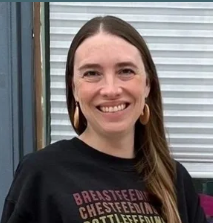 A woman with long brown hair smiling in front of a blue wall and window, wearing a black shirt.