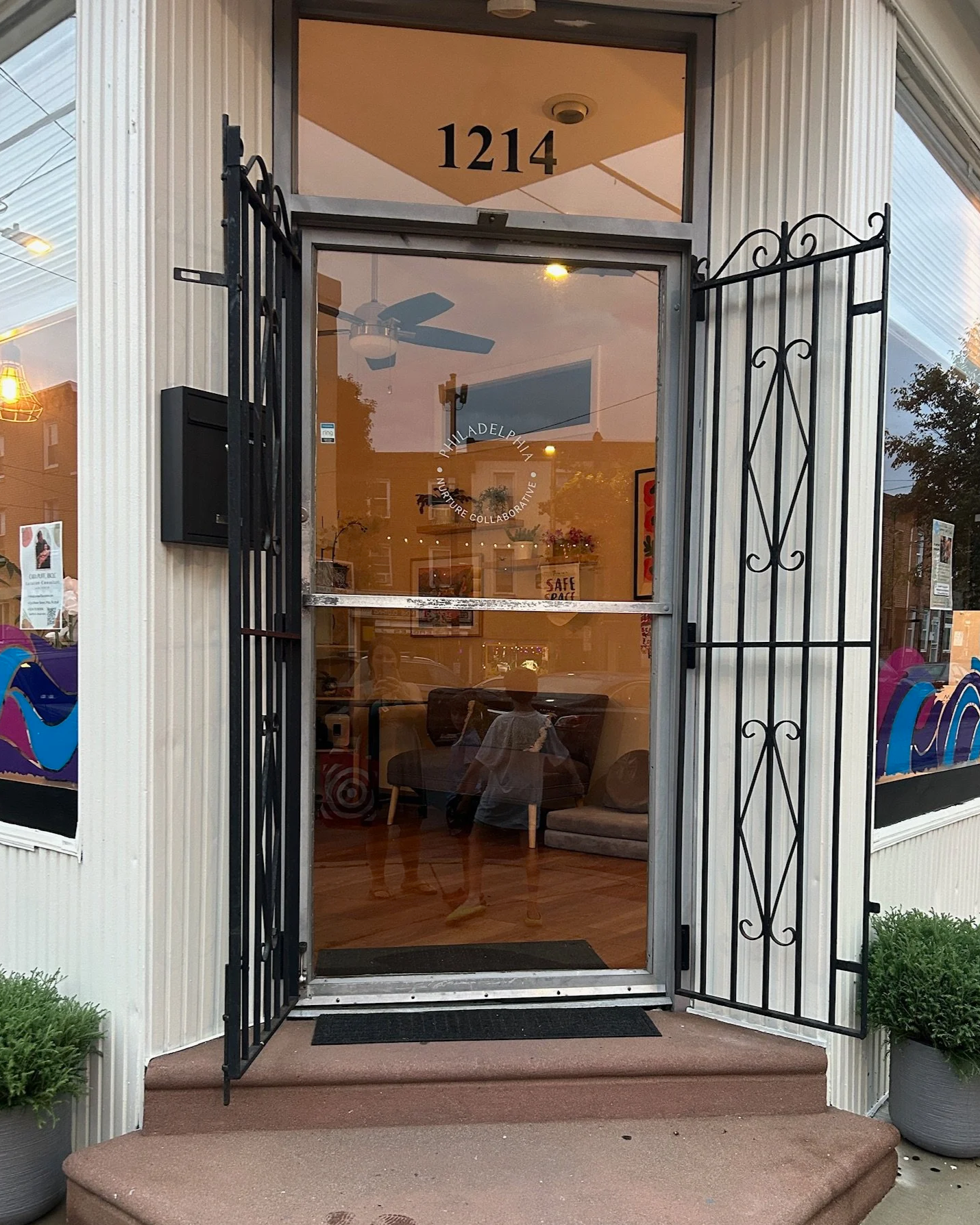 Front entrance of a building with a glass door and window, with the number 1214 displayed above. The door has a black wrought iron security gate open on the right side. Inside, a ceiling fan and some artwork are visible. Two people are reflected on the glass door.