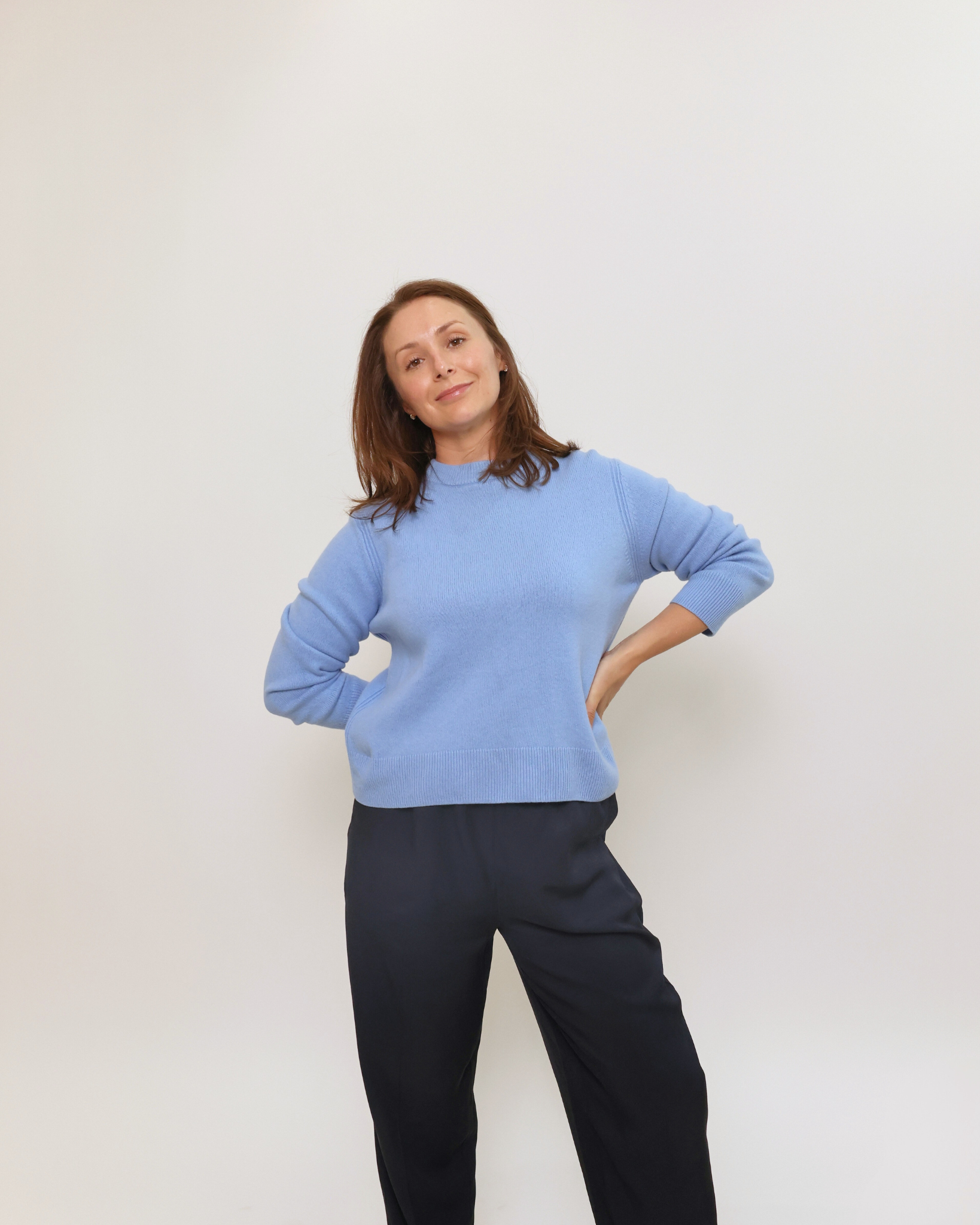 Charlie Perry; a facialist with brown hair wearing a light blue sweater and black pants poses with her hand on her hip against a plain white wall.