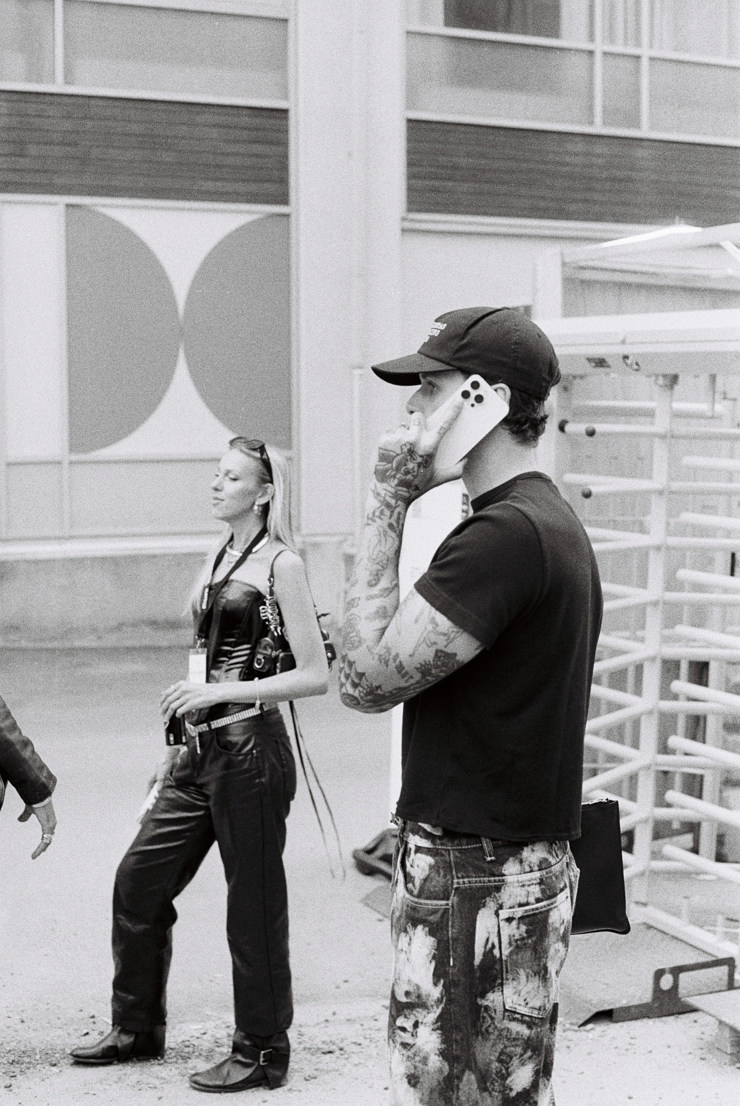 A man with tattoos wearing a cap talking on his cell phone while walking in an urban environment. A woman with sunglasses hanging from her top and a camera around her neck walks nearby. The background includes a modern structure with geometric patterns.