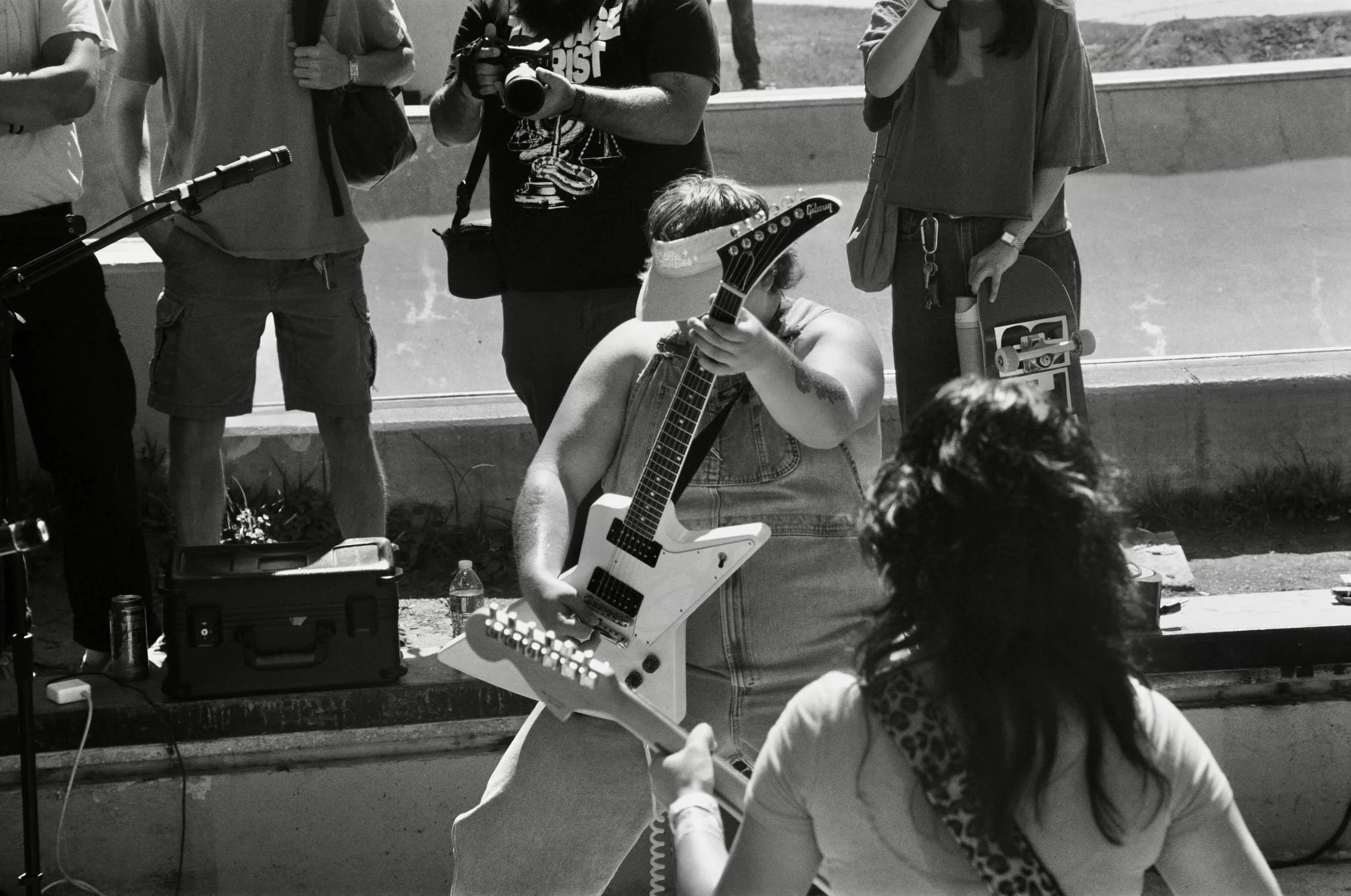 A person holding an Gibson electric guitar, surrounded by people, some with cameras and skateboards, outdoors near a concrete wall.