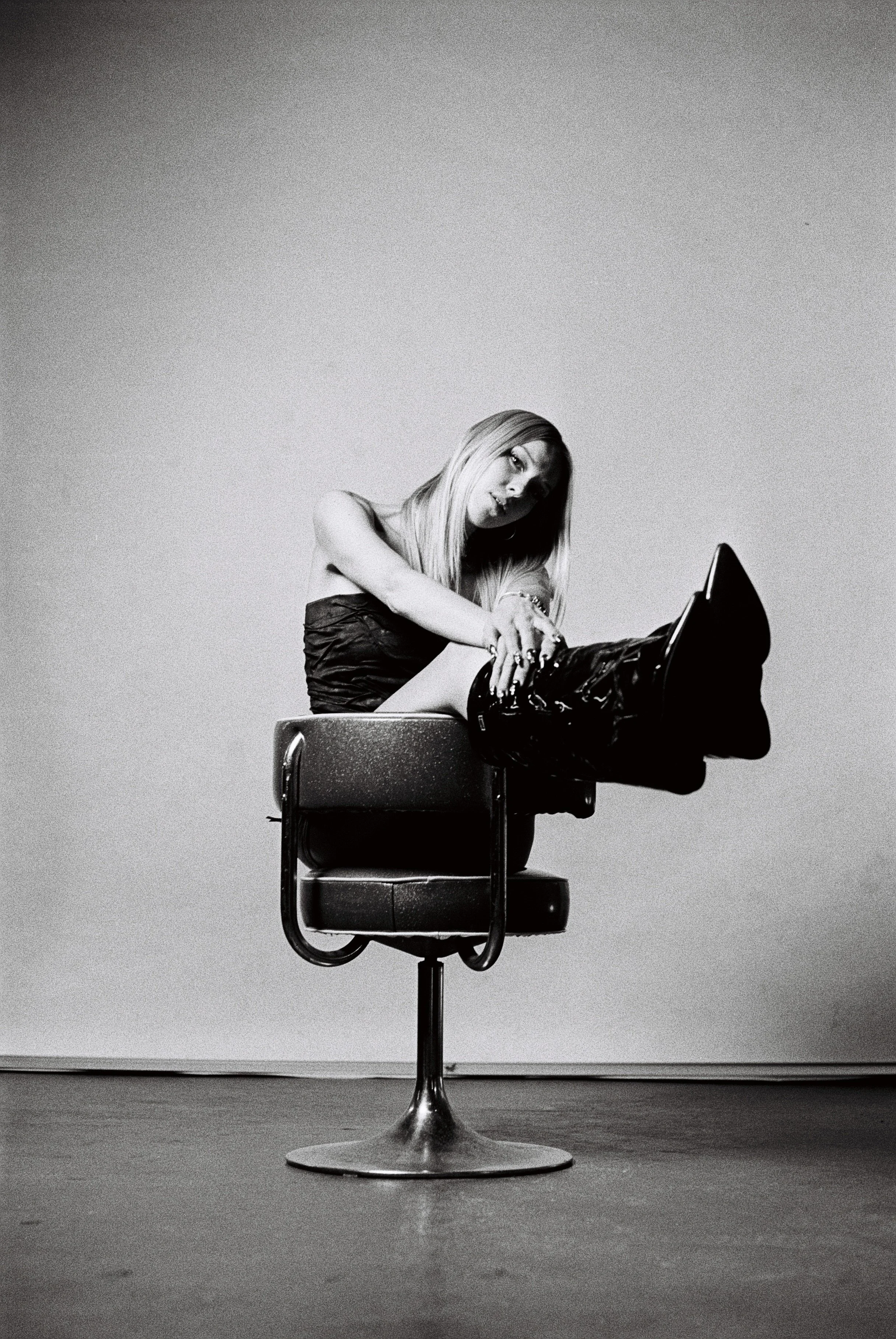 Black and white photo of a woman sitting on an office chair, reaching down to adjust her shoe, against a plain background.
