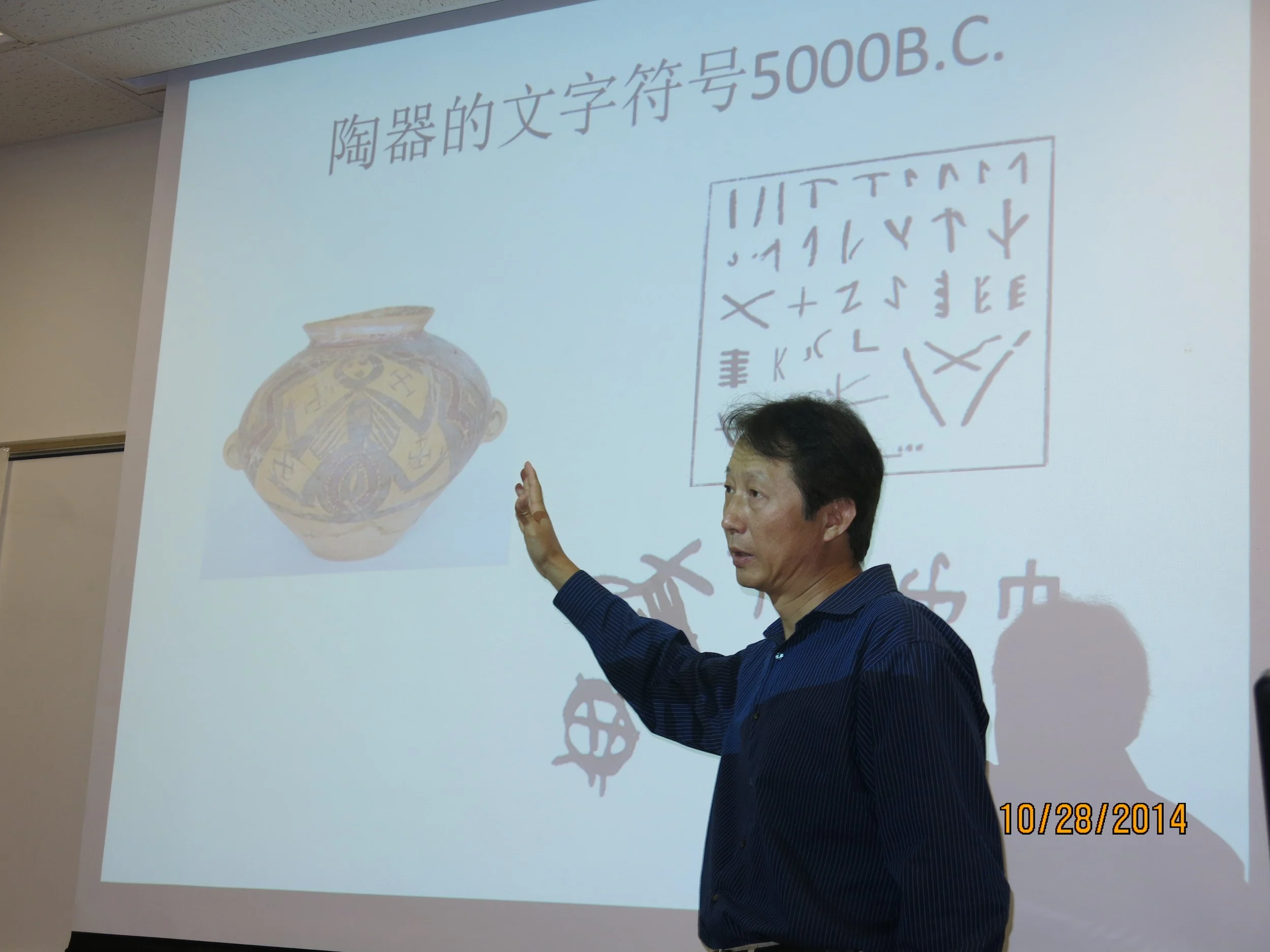 A man in a blue shirt is pointing at a projector screen displaying Chinese characters, a diagram of a decorated vase, and examples of symbols or runes, with the date 10/28/2014 in the lower right corner.