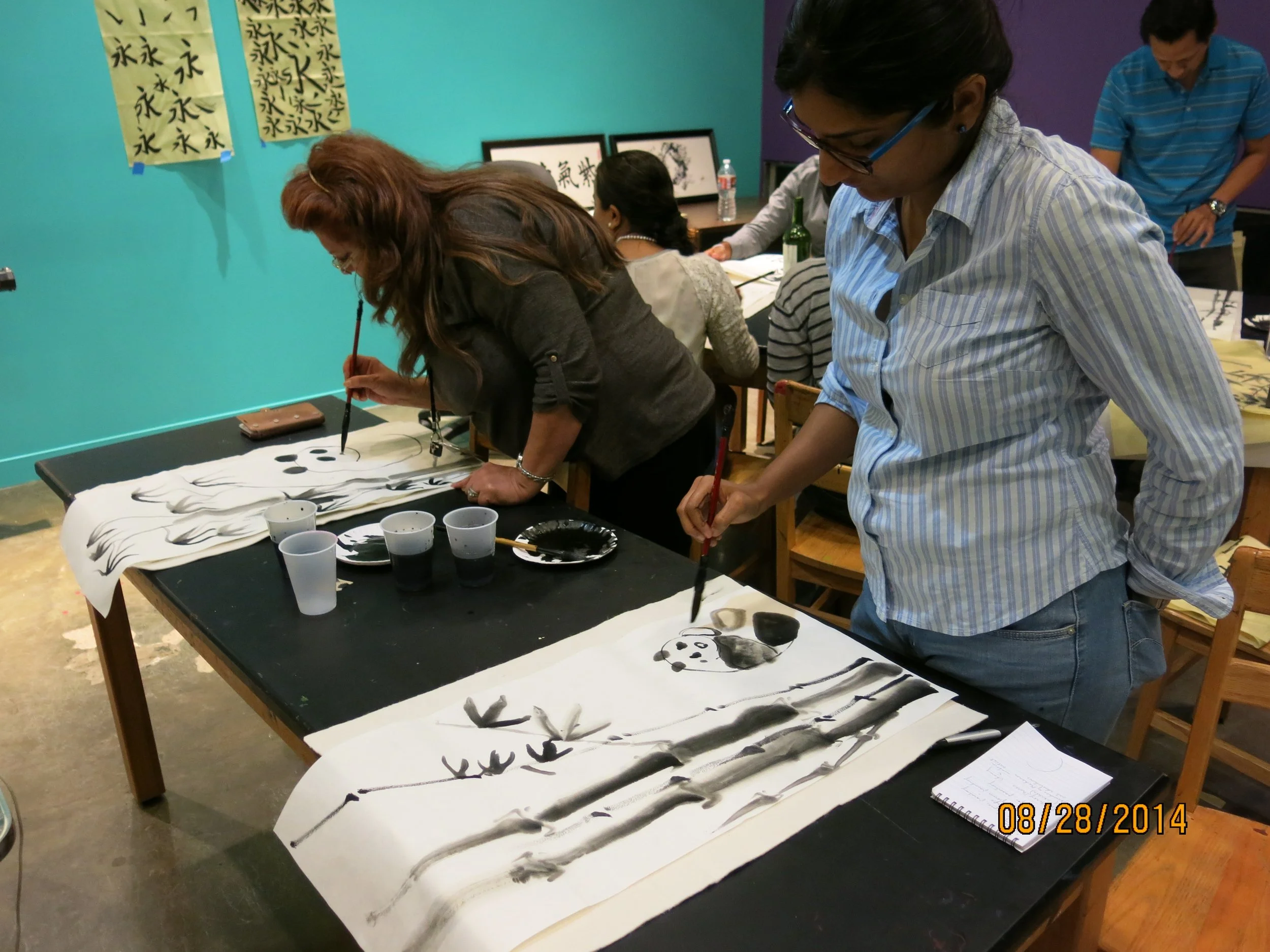 People practicing traditional Asian ink brush painting with black ink on long white paper, displaying various brush strokes and characters.