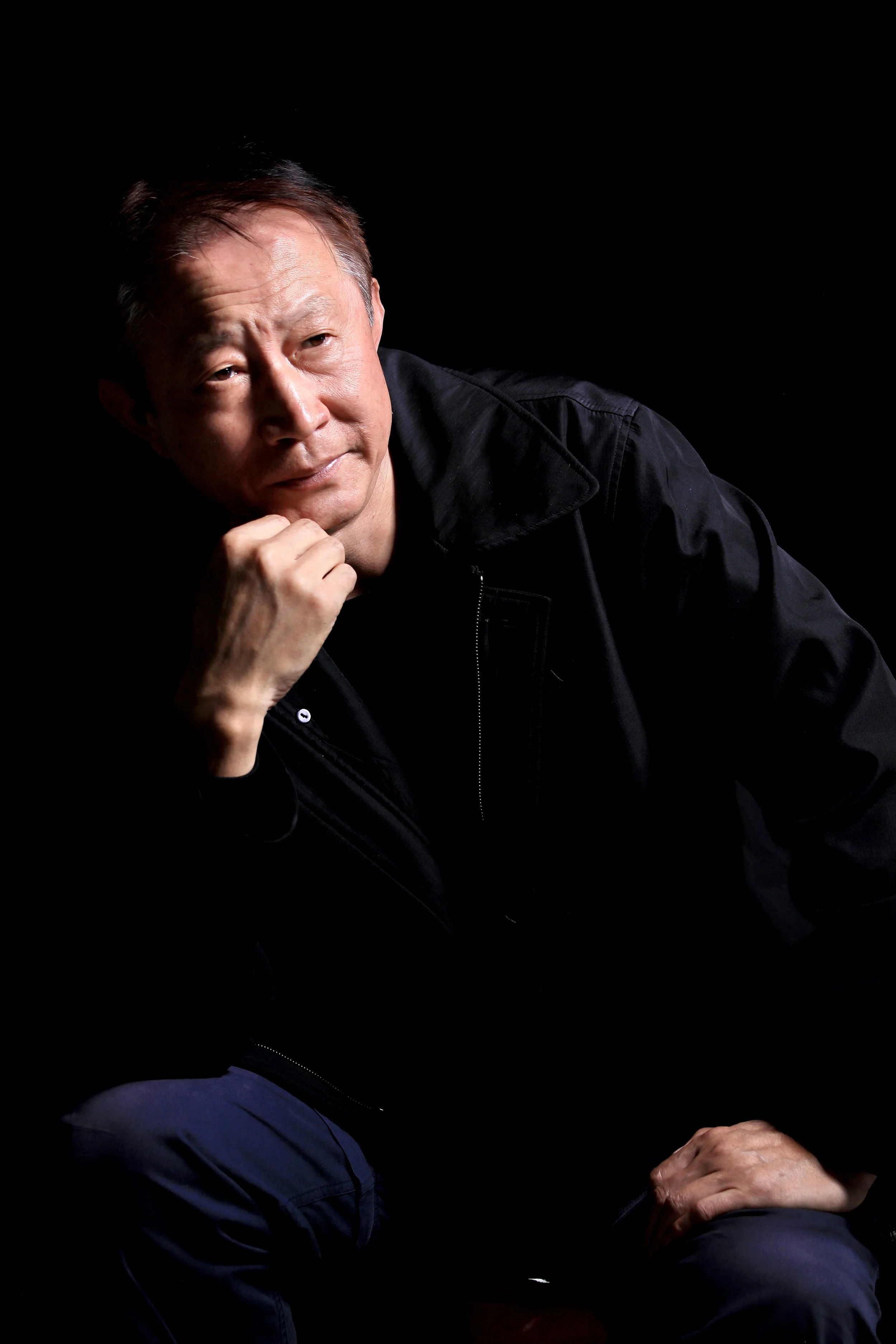 A middle-aged man with short dark hair and a thoughtful expression, resting his chin on his hand against a black background.