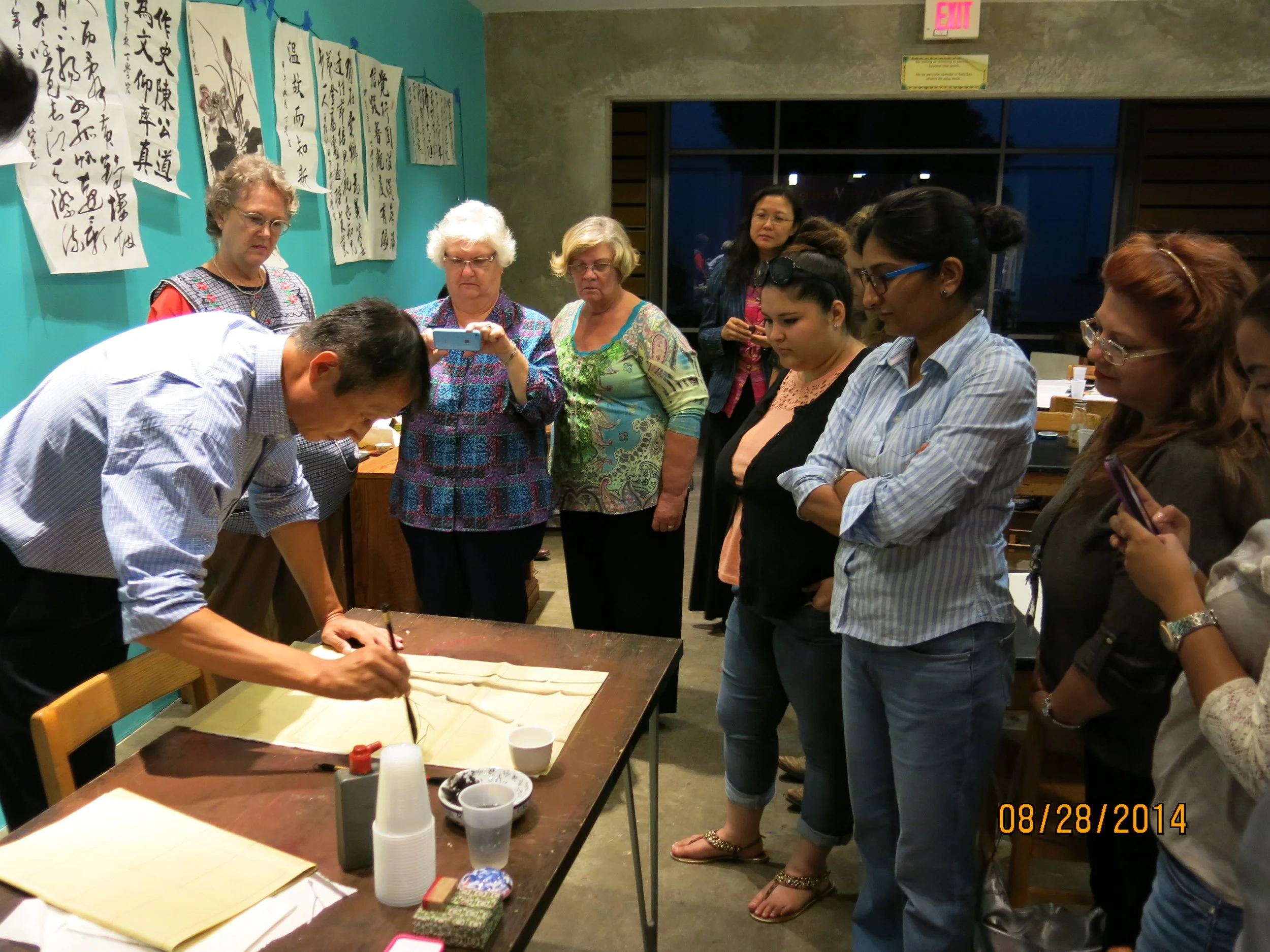 A group of women and a man gathered around a table, watching a man demonstrate a craft or cooking technique. The women are standing, with some holding phones or taking photos. The background features Japanese calligraphy artwork on the wall and a window at night.