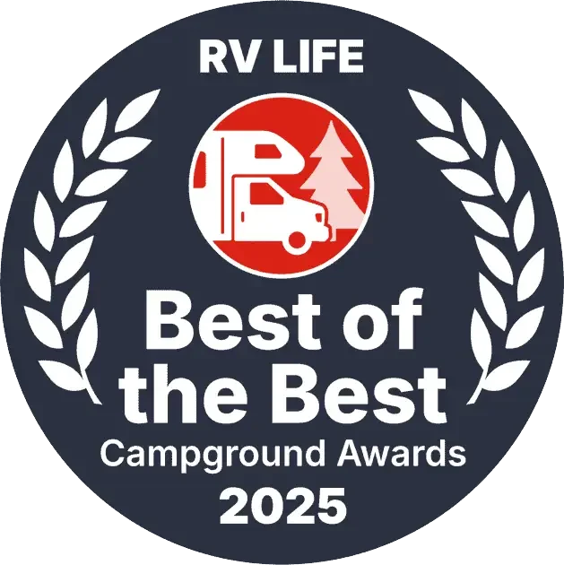 A circular award badge with a dark blue background, white laurel leaves on each side, and text that reads "RV LIFE Best of the Best Campground Awards 2025." In the center, there is a red and white icon of a camper van with a pine tree behind it.