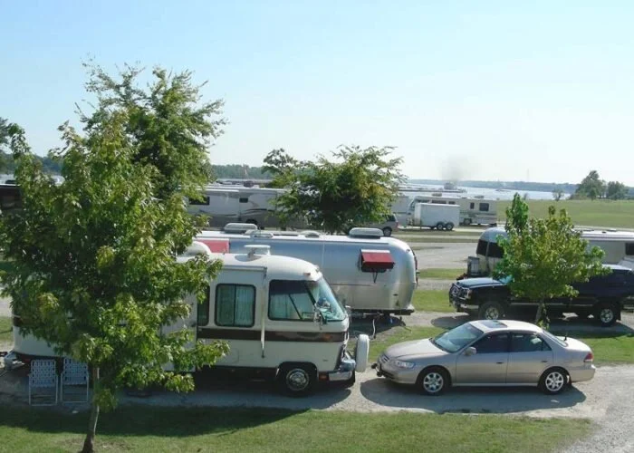 A parking area with motorhomes, cars, and trees, with a body of water in the background.