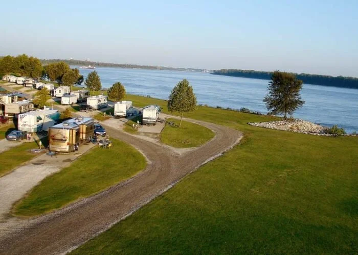 Aerial view of a campground with RVs near a large river, with green grass, trees, and a gravel road winding through the area.