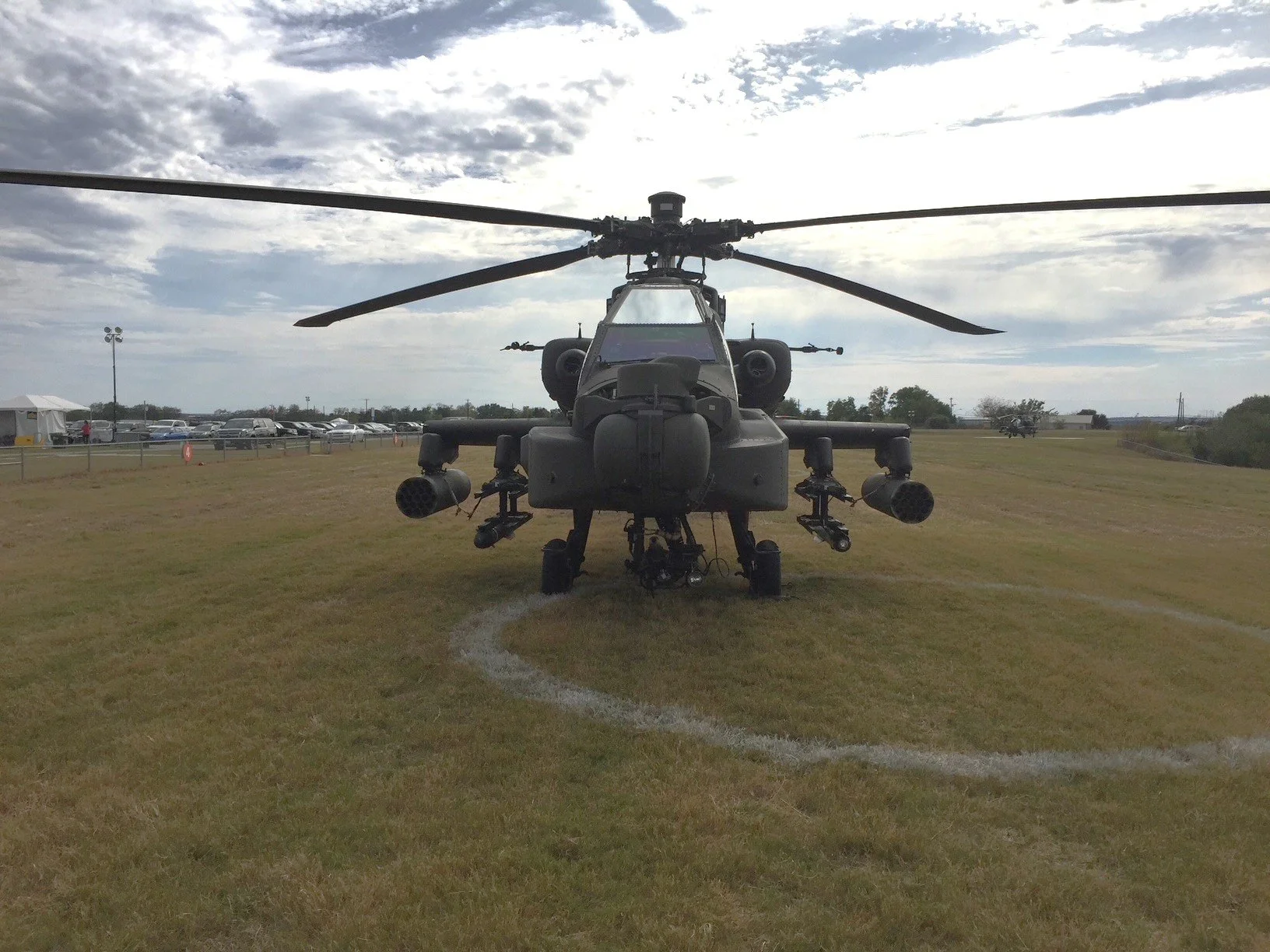 A military attack helicopter displayed on a grassy field at an airshow with cars and tents in the background.