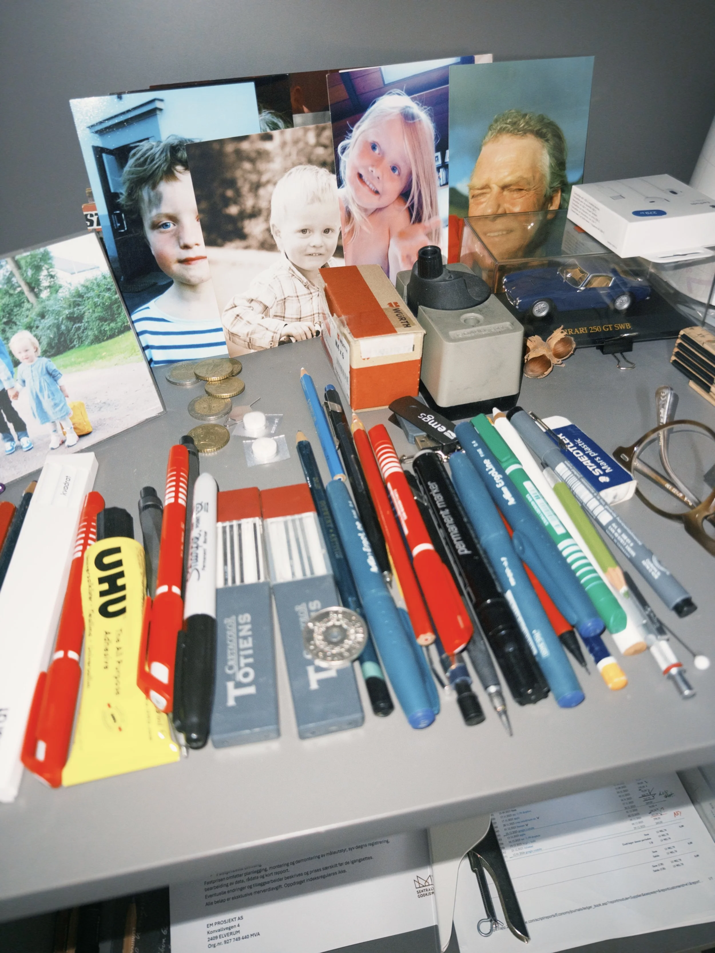 Bilde av en pult med flere fotografier av barn og voksne, forskjellige penner, blyanter, små objekter, et game-tilbehør som en bilmodell, en brusboks og papirarbeid.