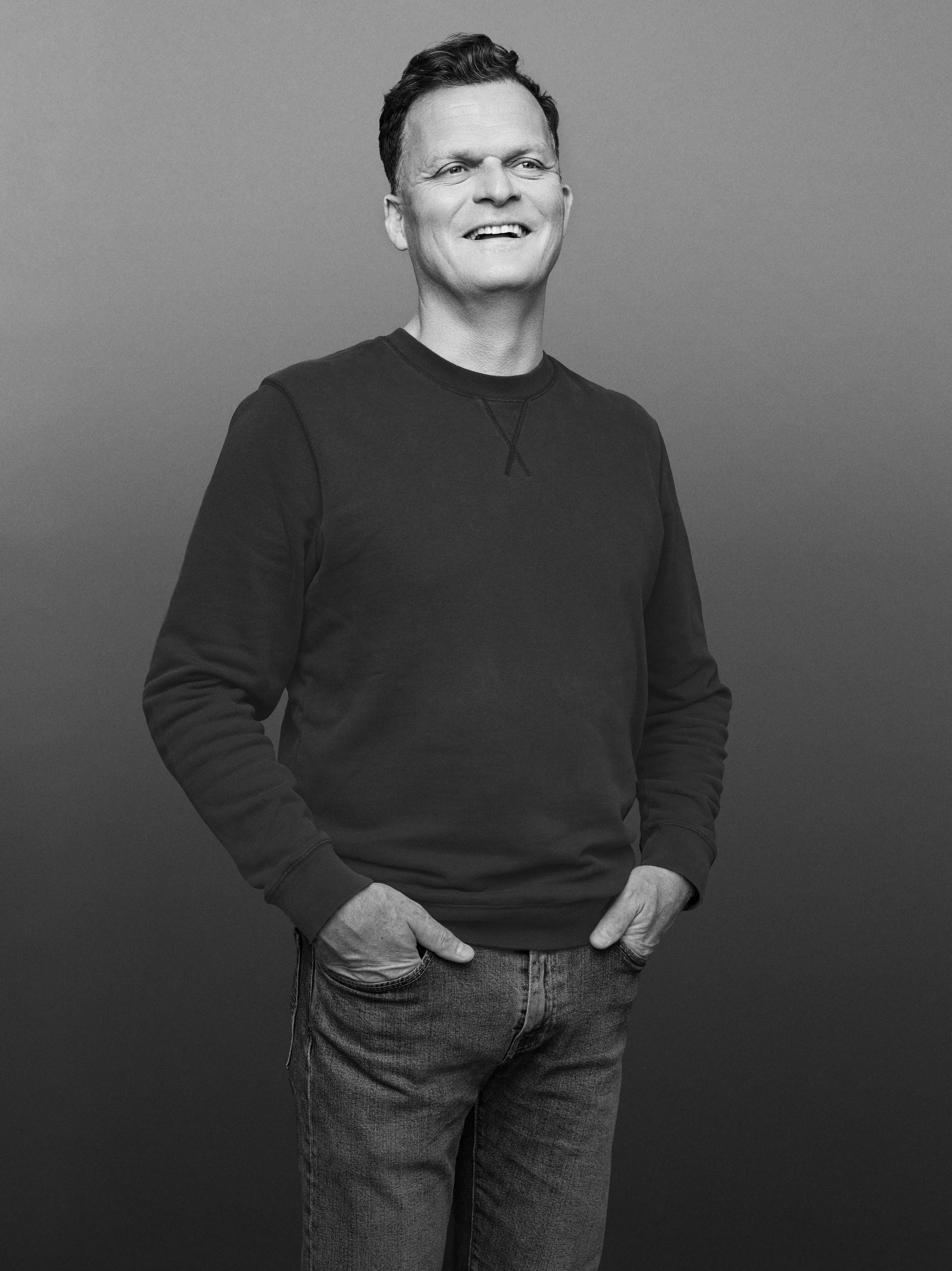 Et svart-hvitt portrett av en mann som smiler, kledd i en svart genser og jeans, mot en enkel grå bakgrunn.