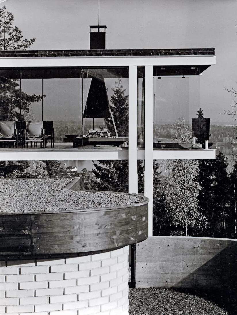 Geir Grung hus. Et moderne hus med store glassvinduer, balkong med møbler og en utedugnad stare peis. Det er trær og landskap i bakgrunnen, og bildet er i svart-hvitt.