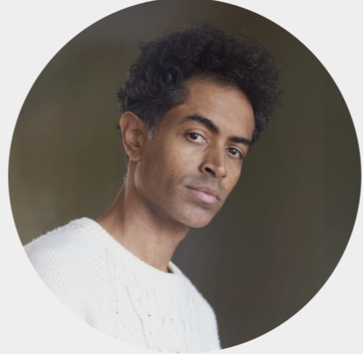 Homem com cabelo cacheado escuro, pele morena clara, usando suéter branco, olhando para a câmera com expressão neutra.