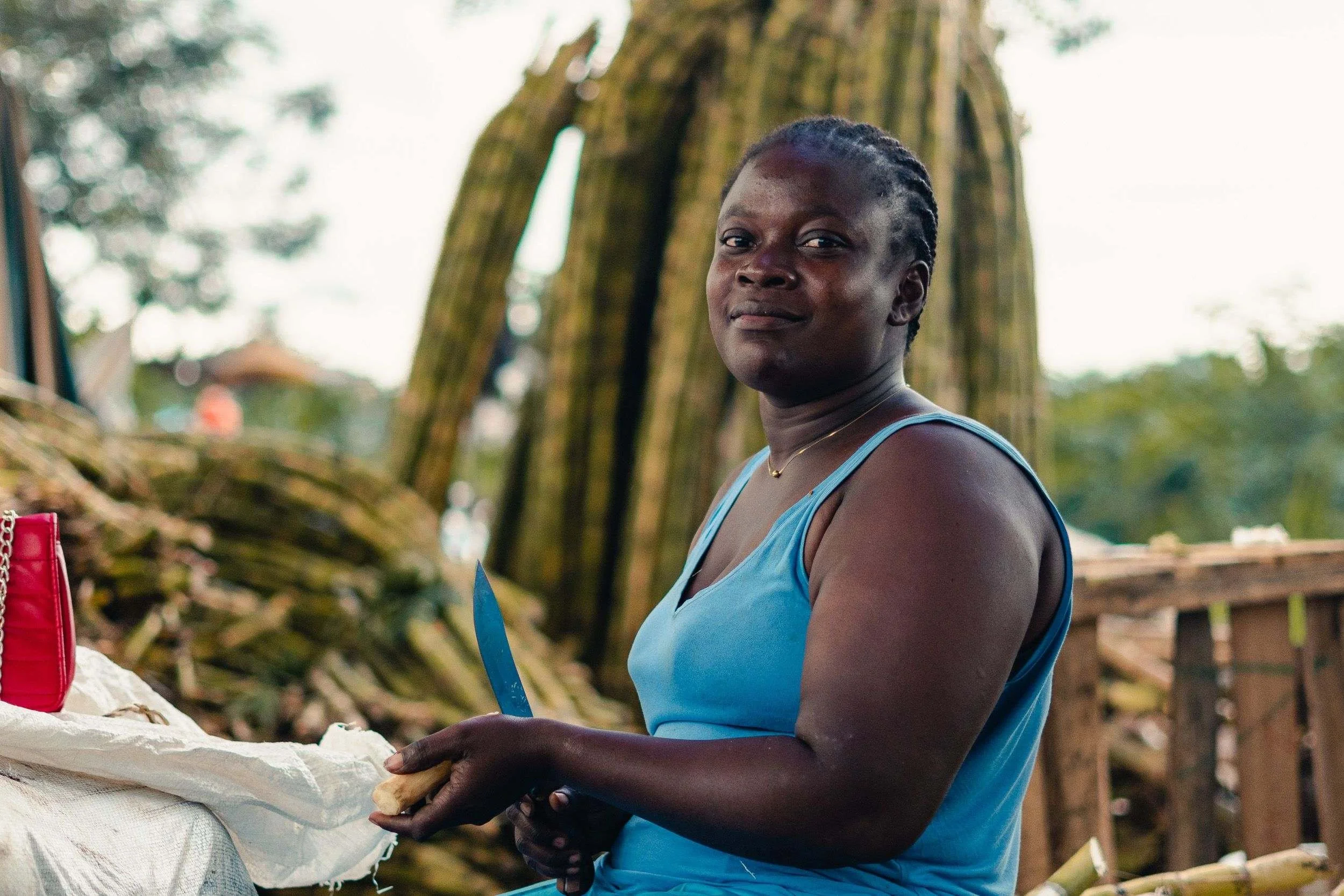 A woman with braided hair, wearing a blue tank top, standing outdoors near a large tree, working with a knife and handling a piece of food.