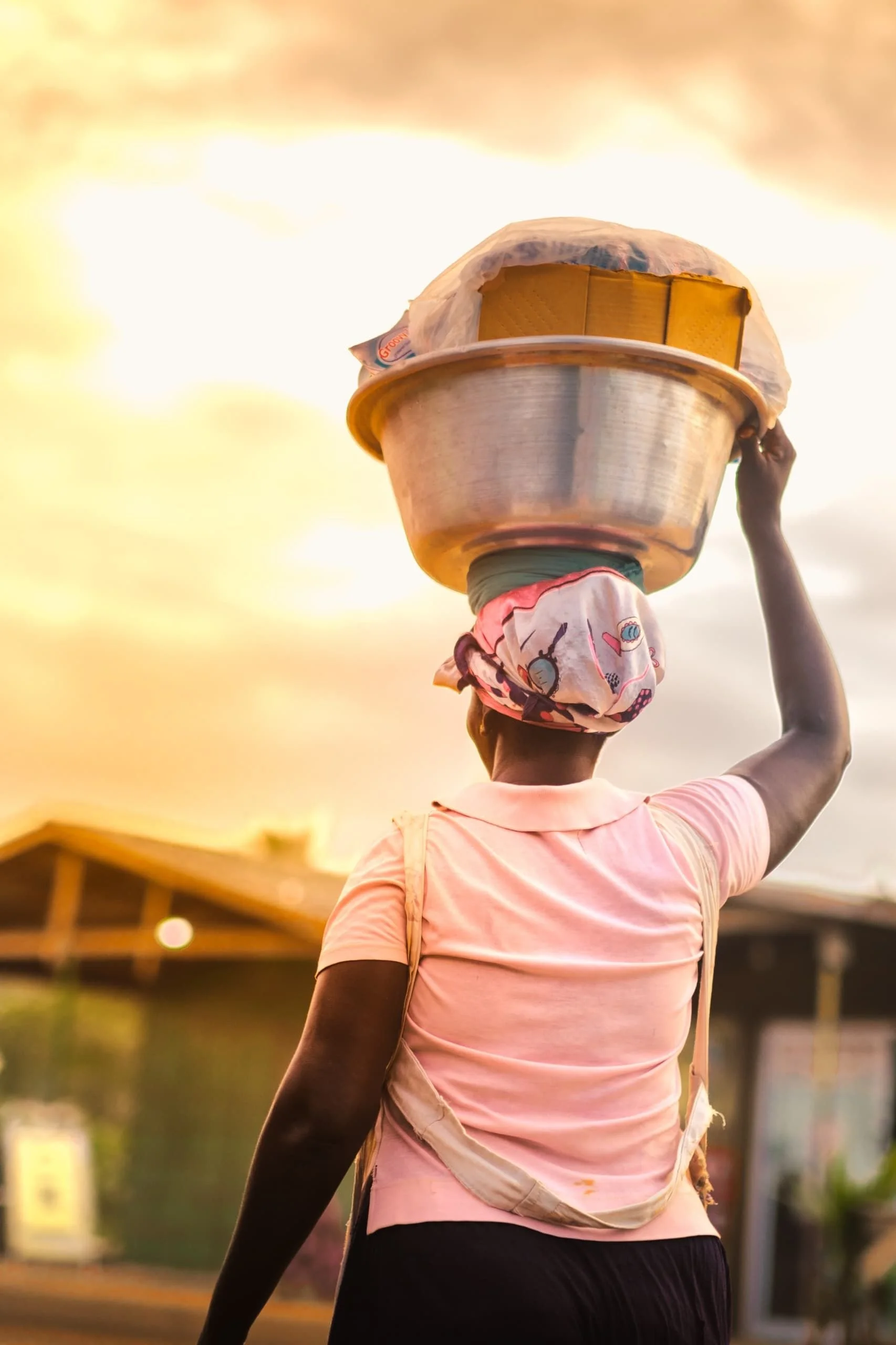 Person carrying a metal bowl on their head, wearing a pink shirt and a colorful headscarf, in an outdoor setting during sunset.