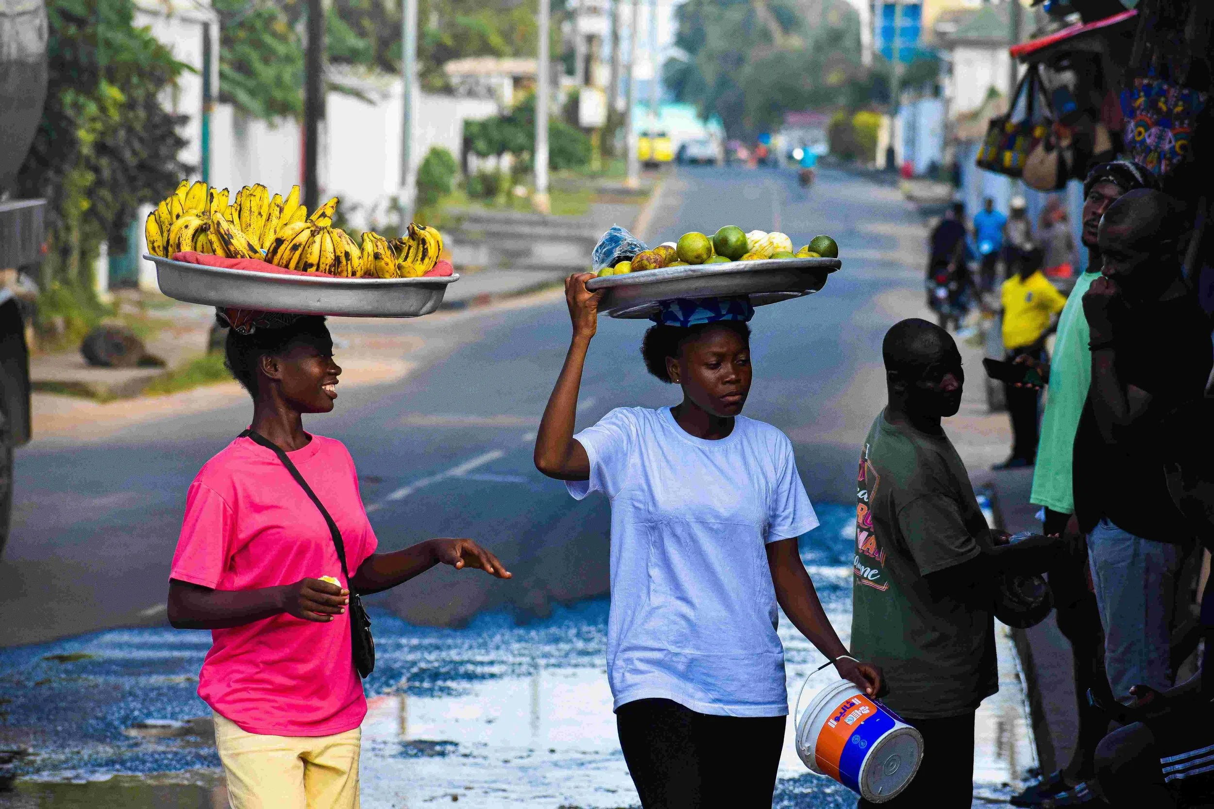 Two women walking on a street, balancing trays of bananas and other fruits on their heads, with other people and vehicles visible in the background.