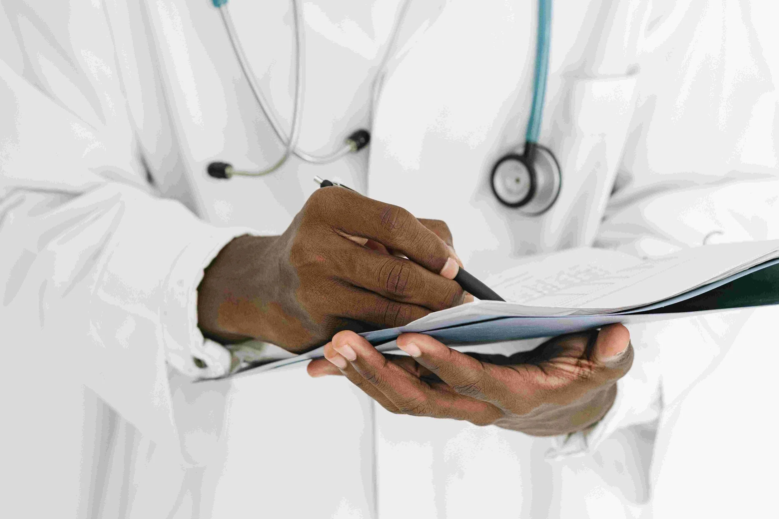 Close-up of a doctor or medical professional in a white coat writing in a clipboard, with a stethoscope hanging around the neck.