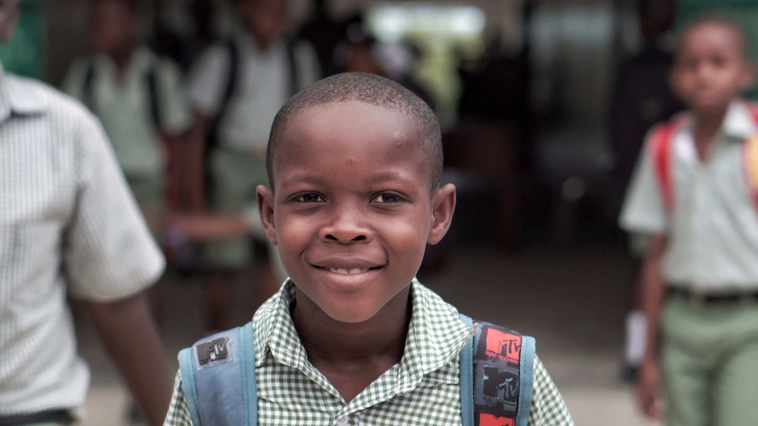Smiling young boy with a backpack in a school uniform, standing among other students in a school corridor.