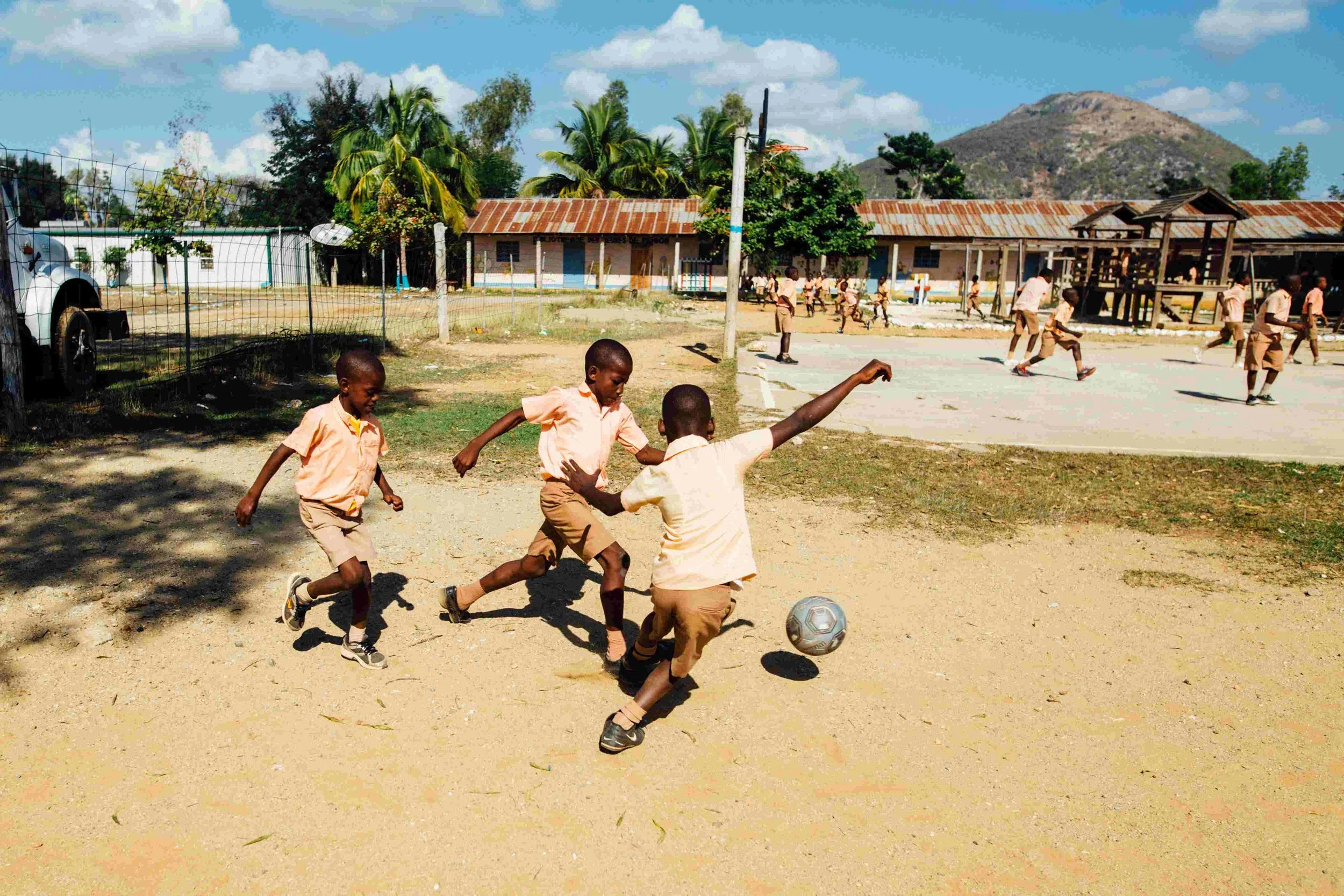 Children playing soccer on a dirt lot outside a school building with multiple levels and corrugated metal roof, surrounded by trees and mountains in the background under a blue sky with clouds.