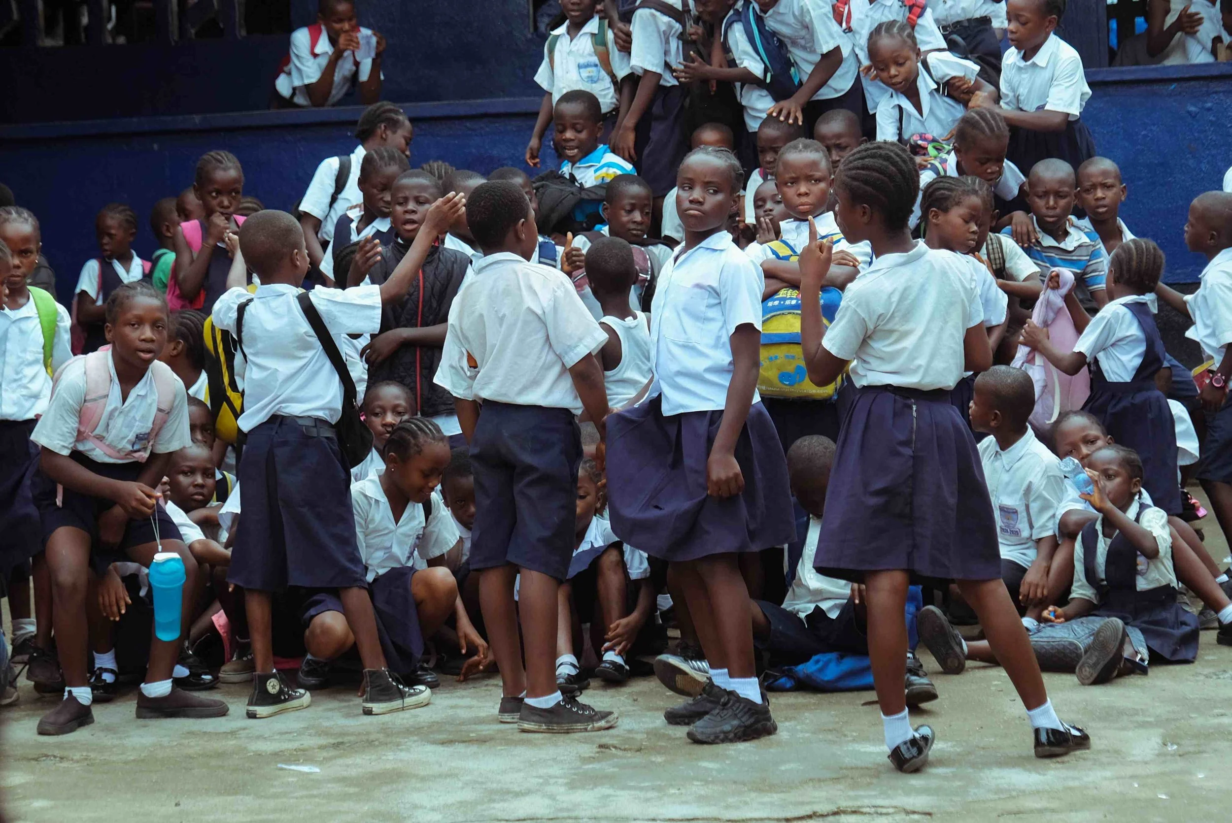 A group of school children gathered outside, some standing and some sitting, wearing white shirts and dark shorts or skirts, with backpacks. A woman is standing among them, giving instructions or talking to the children. The children appear attentive, engaged, and some are exchanging glances or raising their hands.