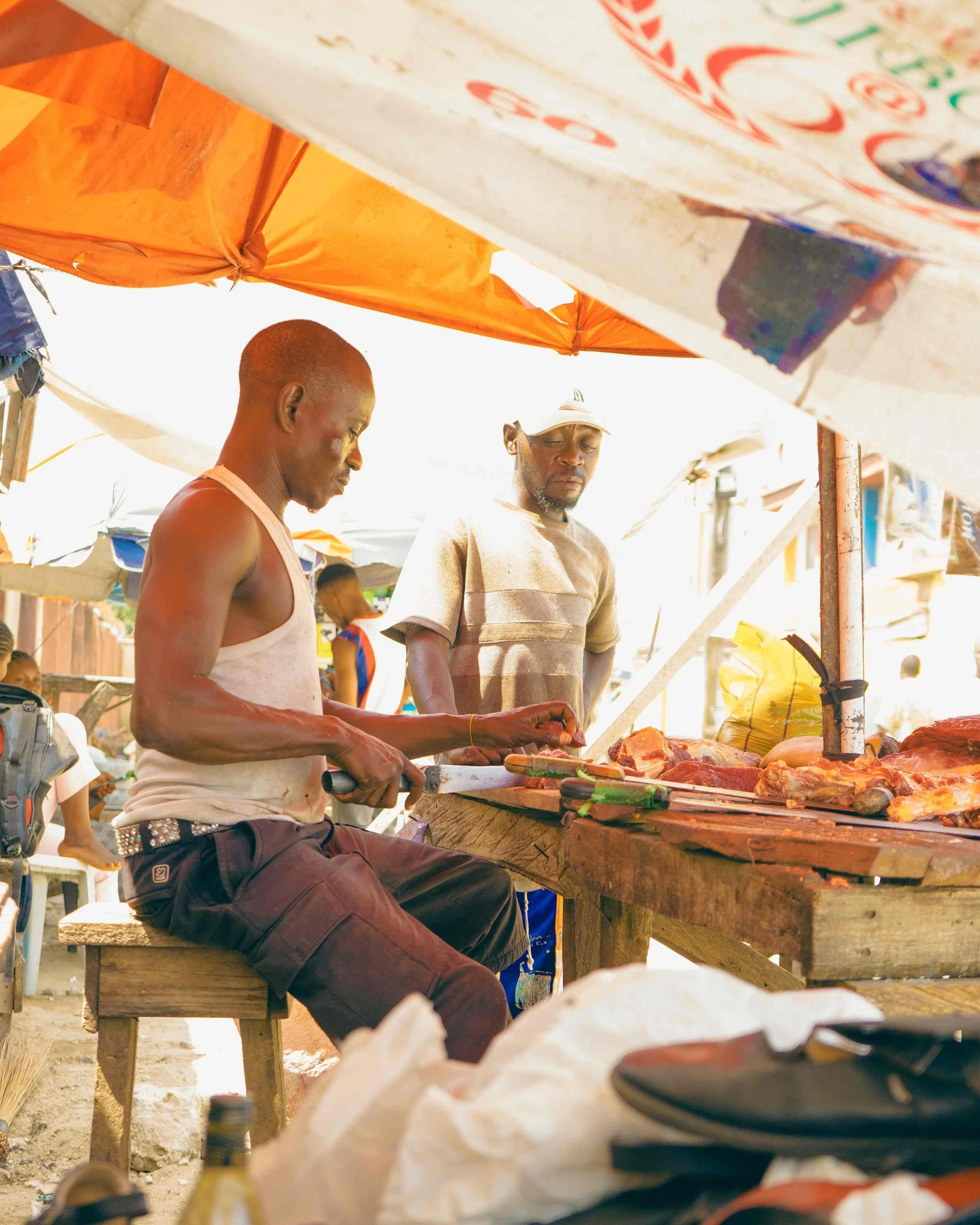 Two men are working at an outdoor market stall selling meat. The man on the left, wearing a sleeveless shirt, is slicing meat, while the man on the right, wearing a cap, looks at the meat. Market umbrellas are visible overhead.