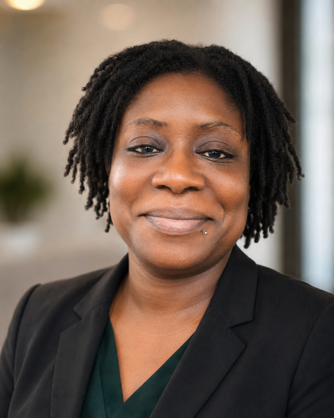 Close-up of a smiling Black woman with short, curly hair, wearing a black blazer and green top, indoors with blurred background.