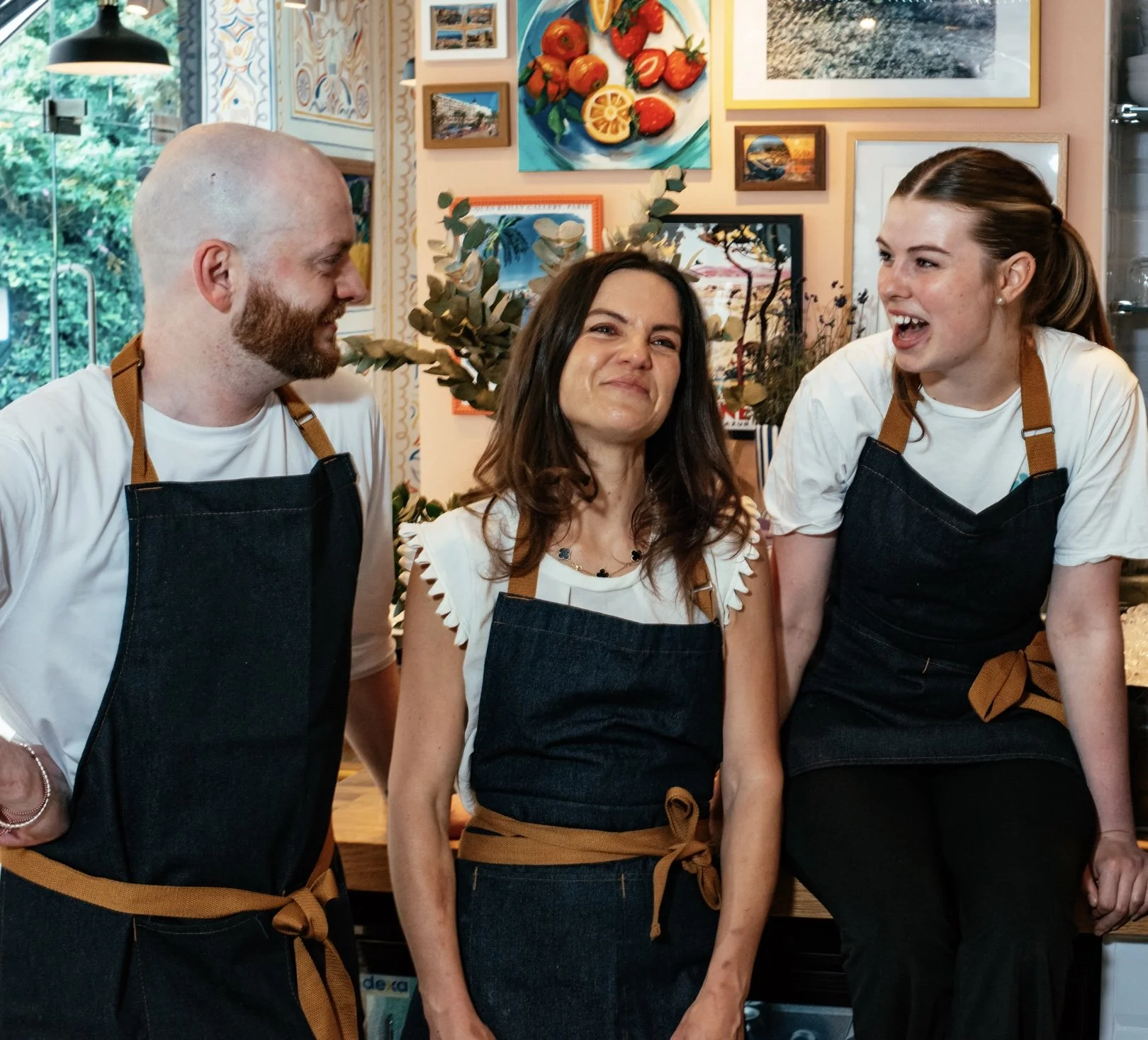 Three people are standing together in a room with colorful artwork on the wall. They are wearing white shirts and black aprons with brown straps, and are smiling or laughing. The room has large windows showing greenery outside.