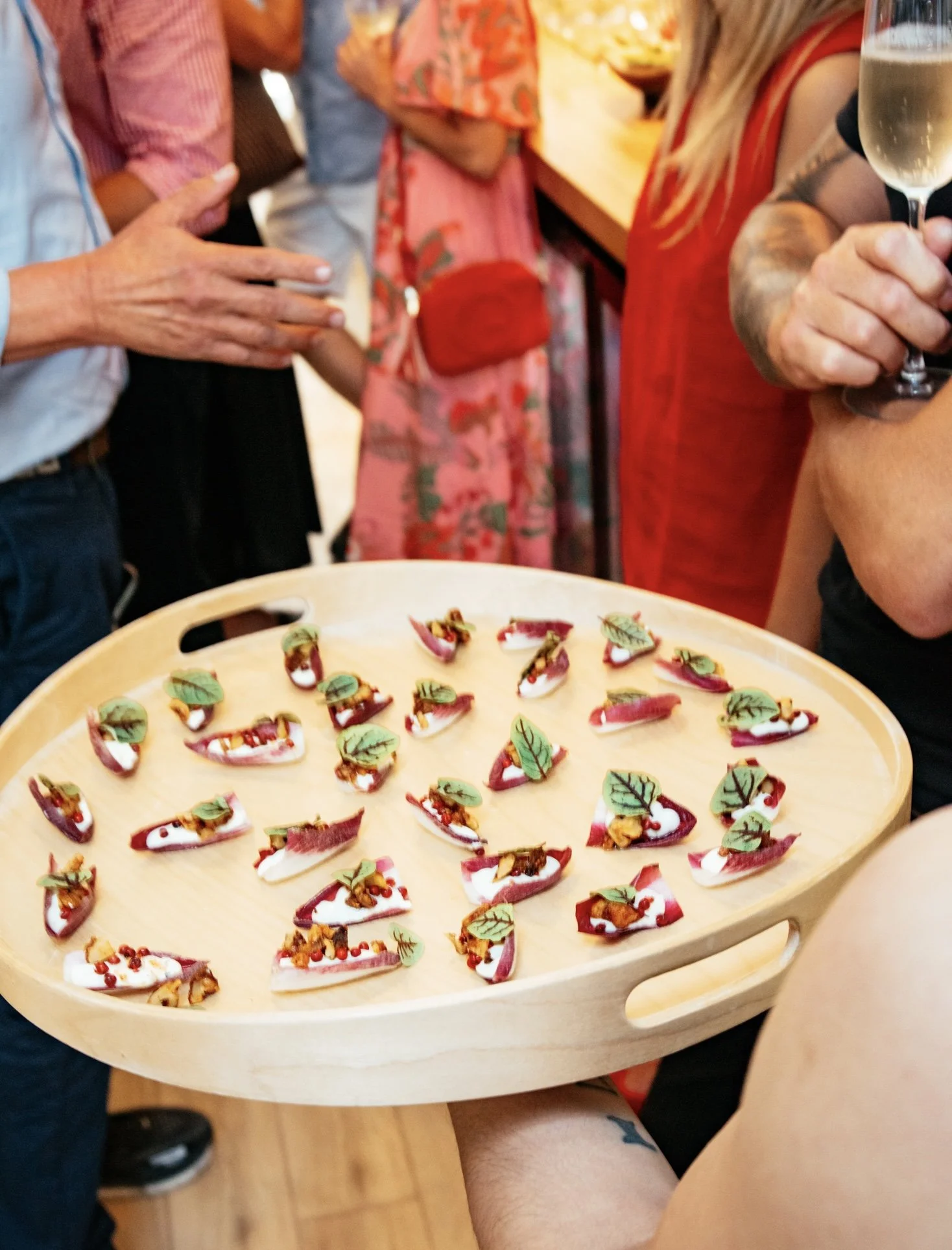 A tray of small, colorful appetizers or tapas with toppings including herbs, nuts, and sauces, being held by a person in a social gathering.