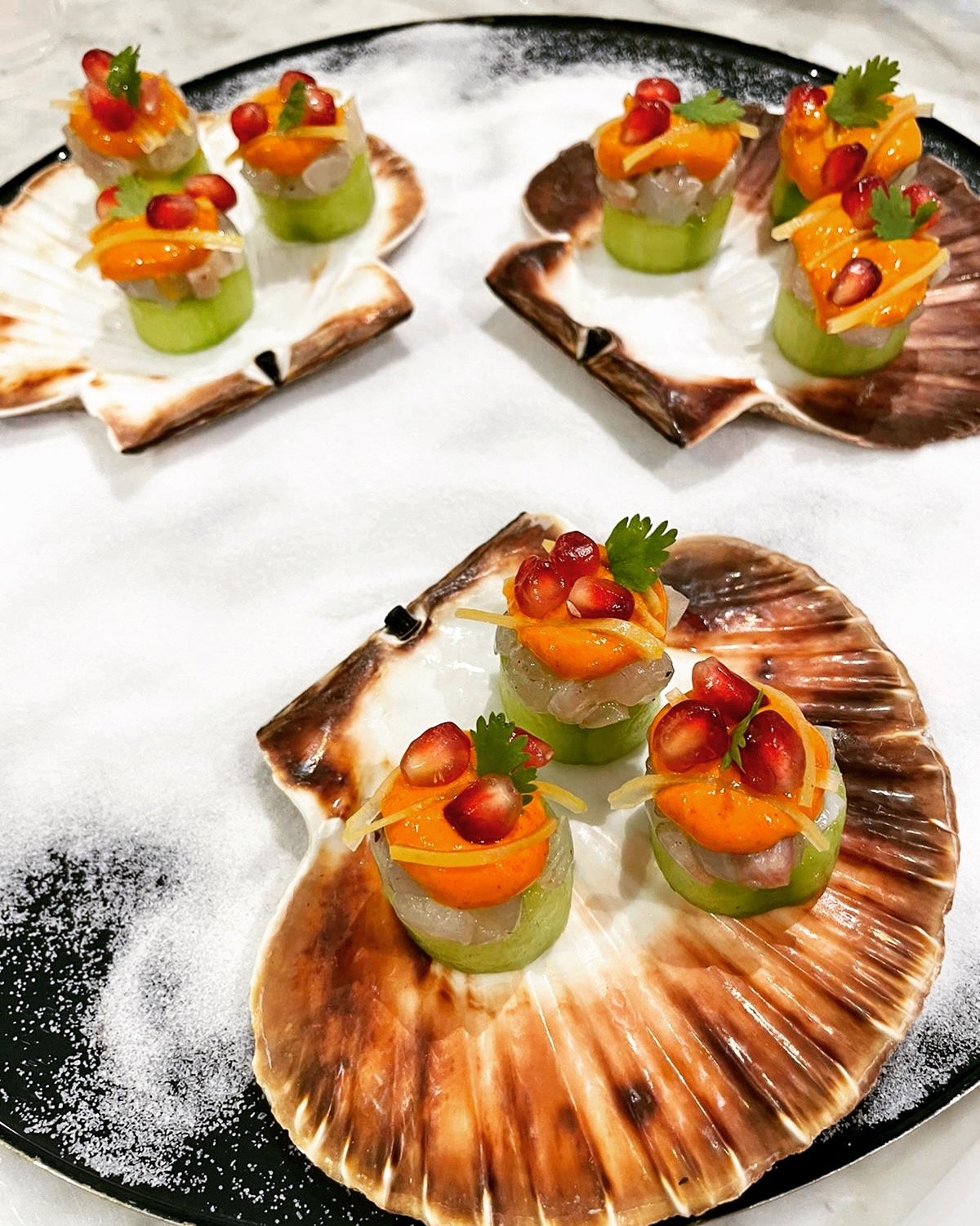 Sea shells with small appetizers containing sliced cucumbers, yellow cheese, diced fish or seafood, pomegranate seeds, and garnished with cilantro, arranged on a black plate.