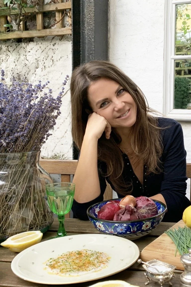 Woman with long brown hair smiling and resting her head on her hand at an outdoor table. The table has a bowl of red onions, a plate of food, lemon wedges, a green glass, and a bouquet of lavender flowers in a glass jar.