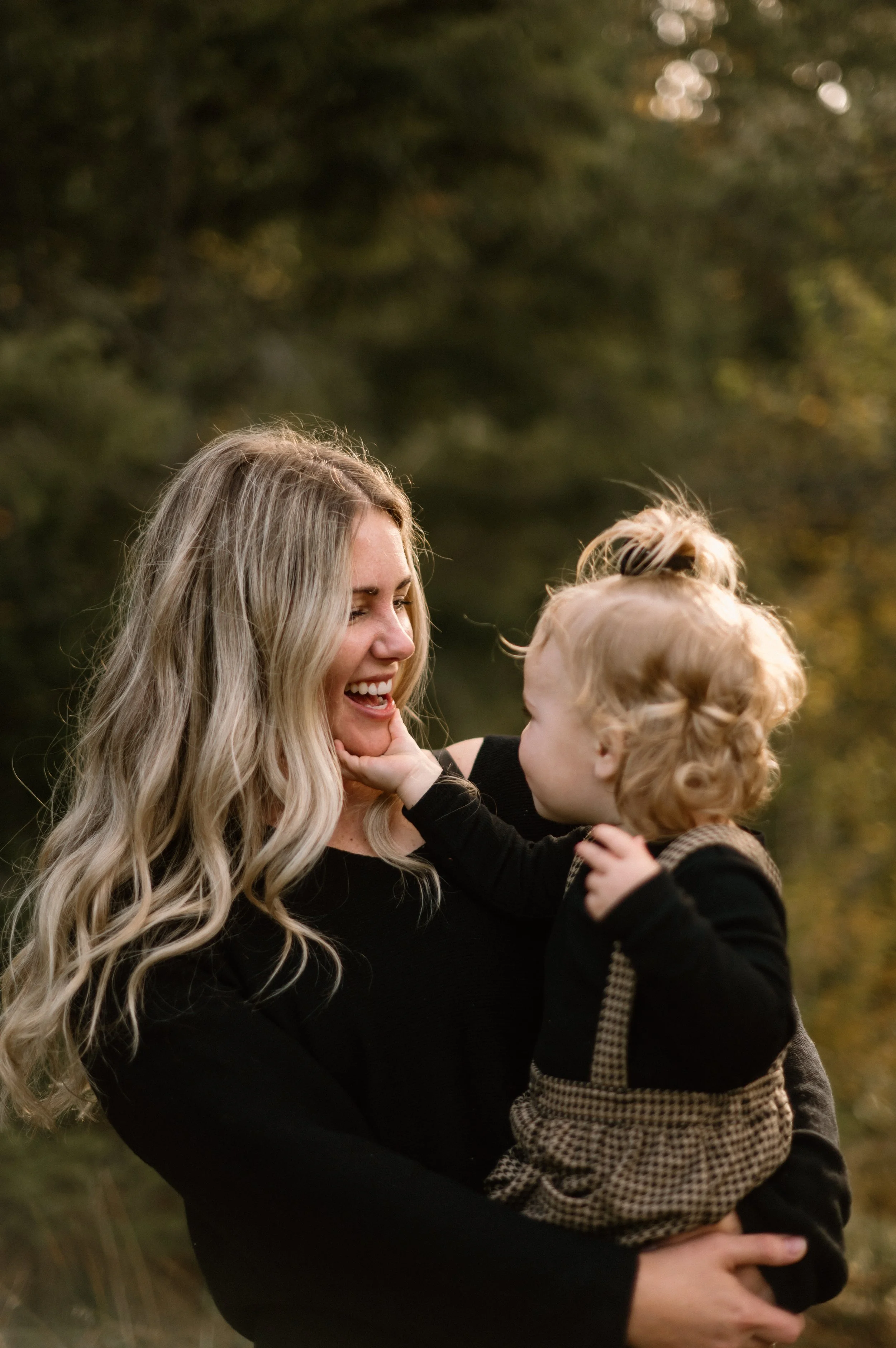 A woman with long blonde hair smiling and holding a young blonde-haired child outdoors.